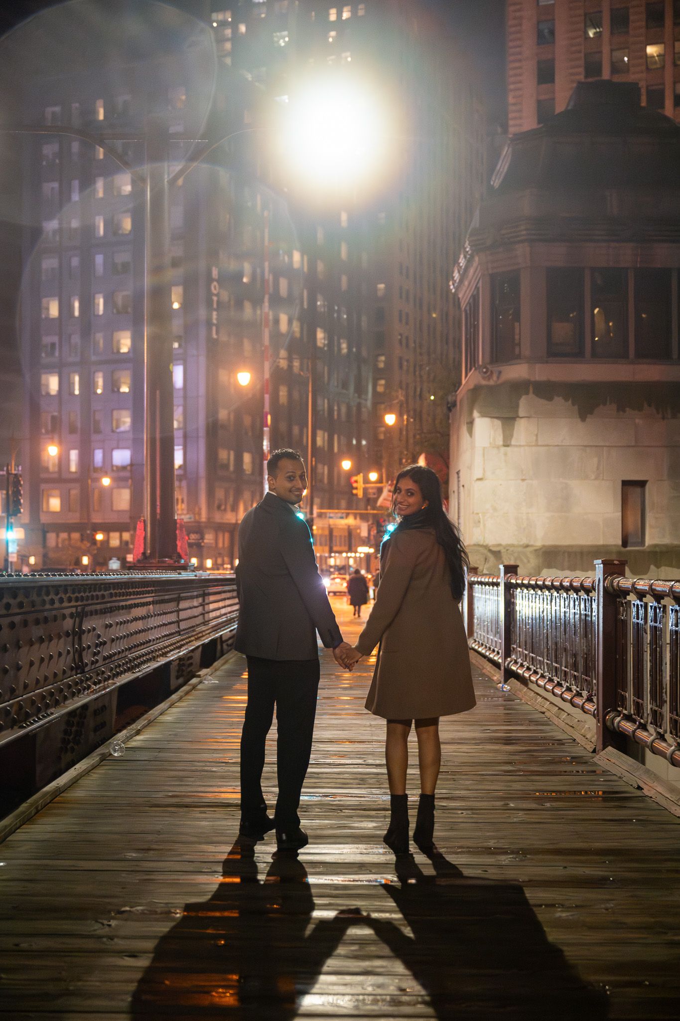 Silhouetted couple holding hands on illuminated bridge at night with city lights in background.