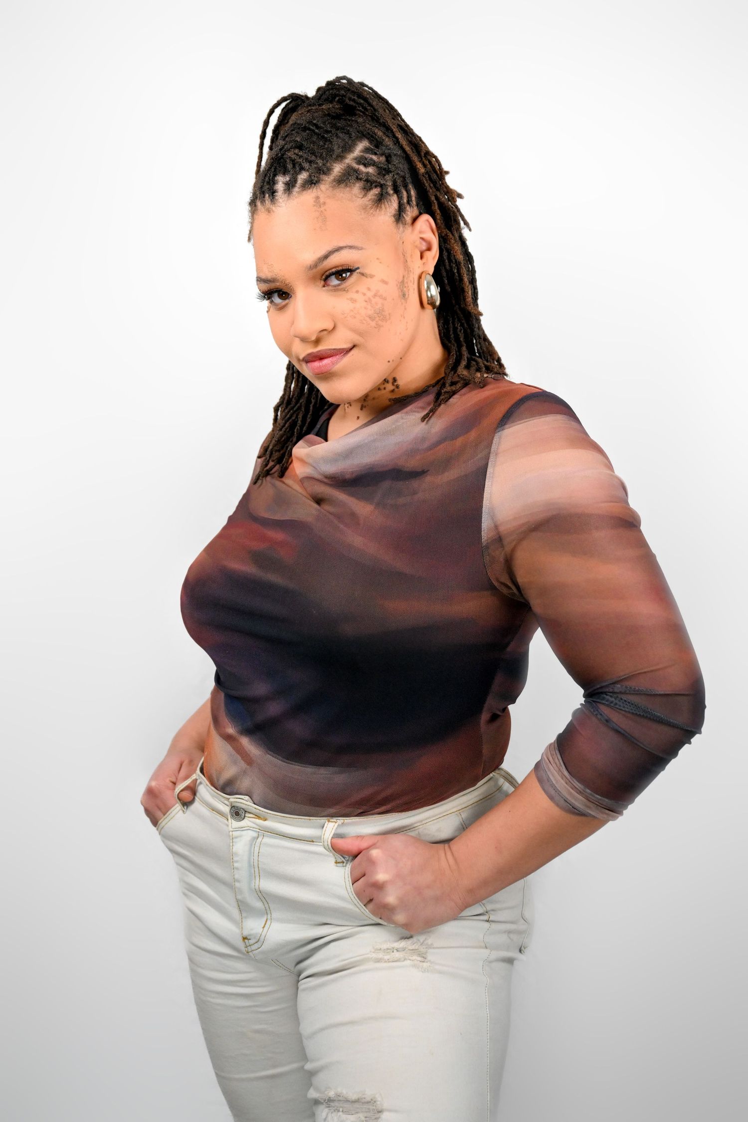 Person in sheer brown top and white pants posing confidently against white background.