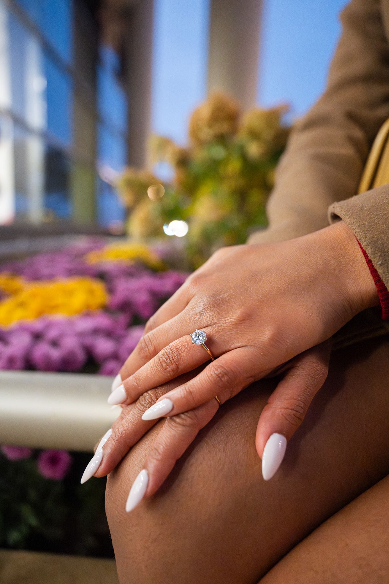White manicured nails and diamond engagement ring displayed with purple flowers in the background.