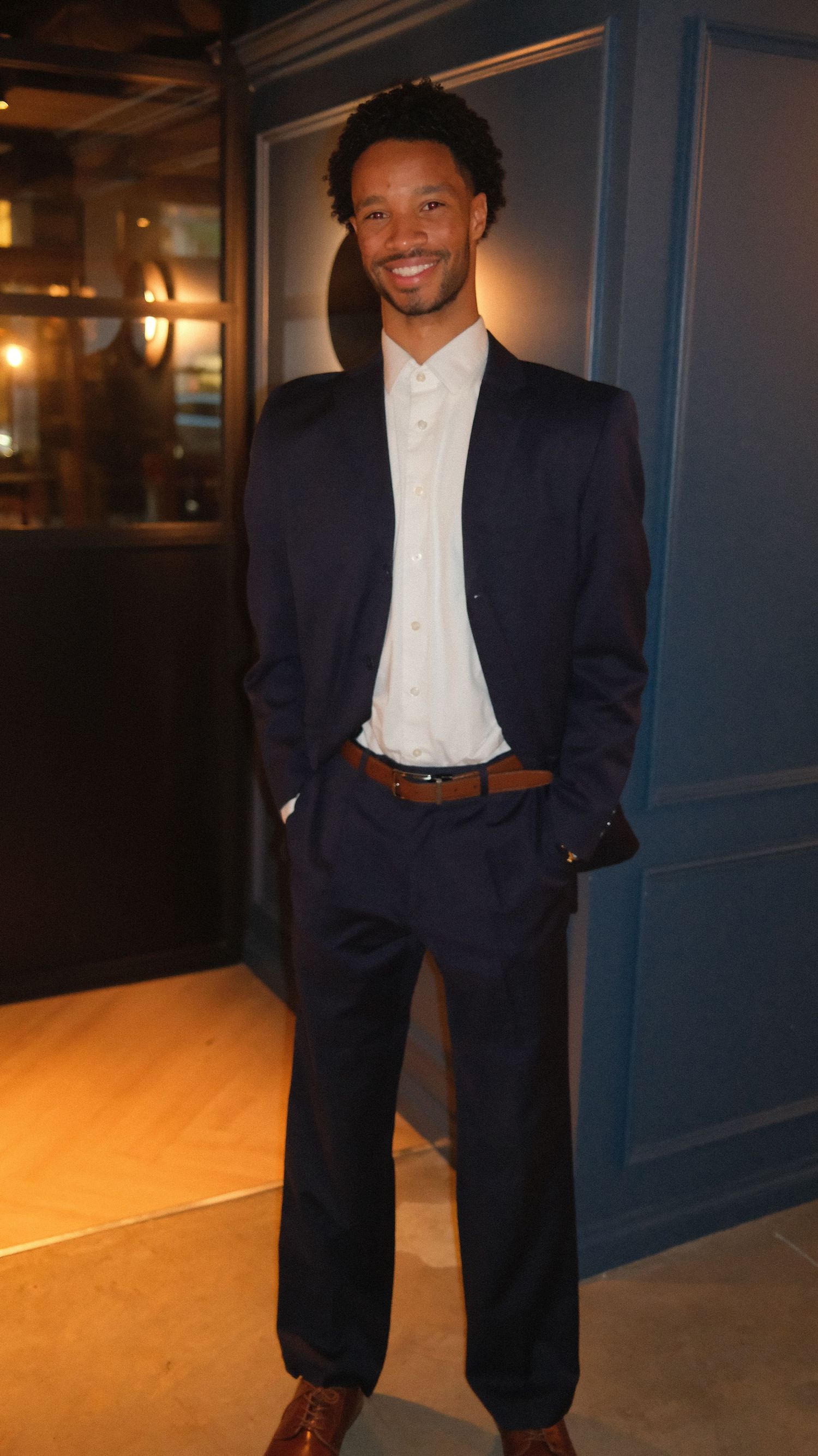 Person wearing a navy blue blazer and white shirt poses in a dimly lit hallway with warm lighting.