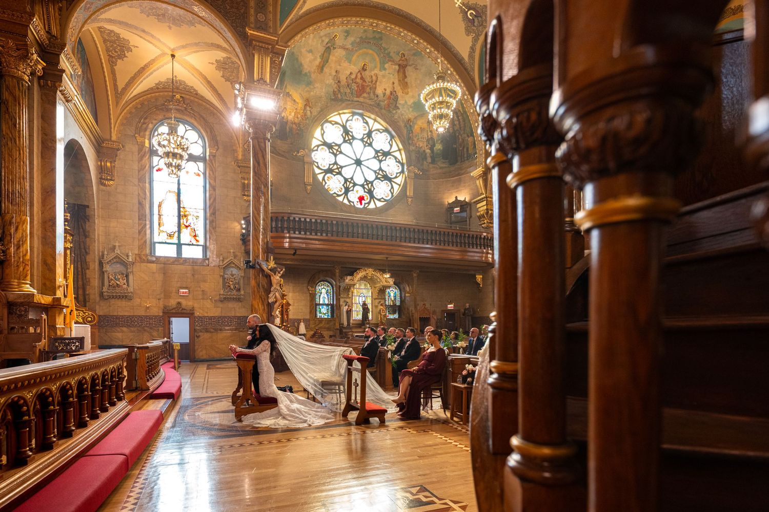 Ornate Gothic church interior with wooden pews, stone columns, and stained glass windows illuminated by natural light.