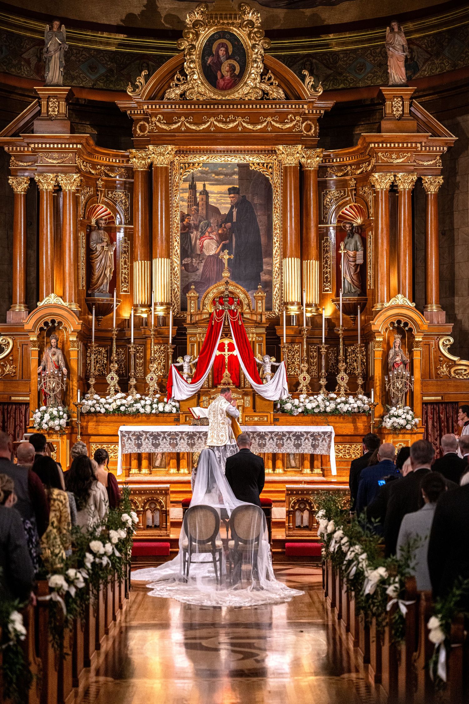 A wedding ceremony in a grand ornate cathedral with gold baroque architecture, white floral decorations, and candlelight.