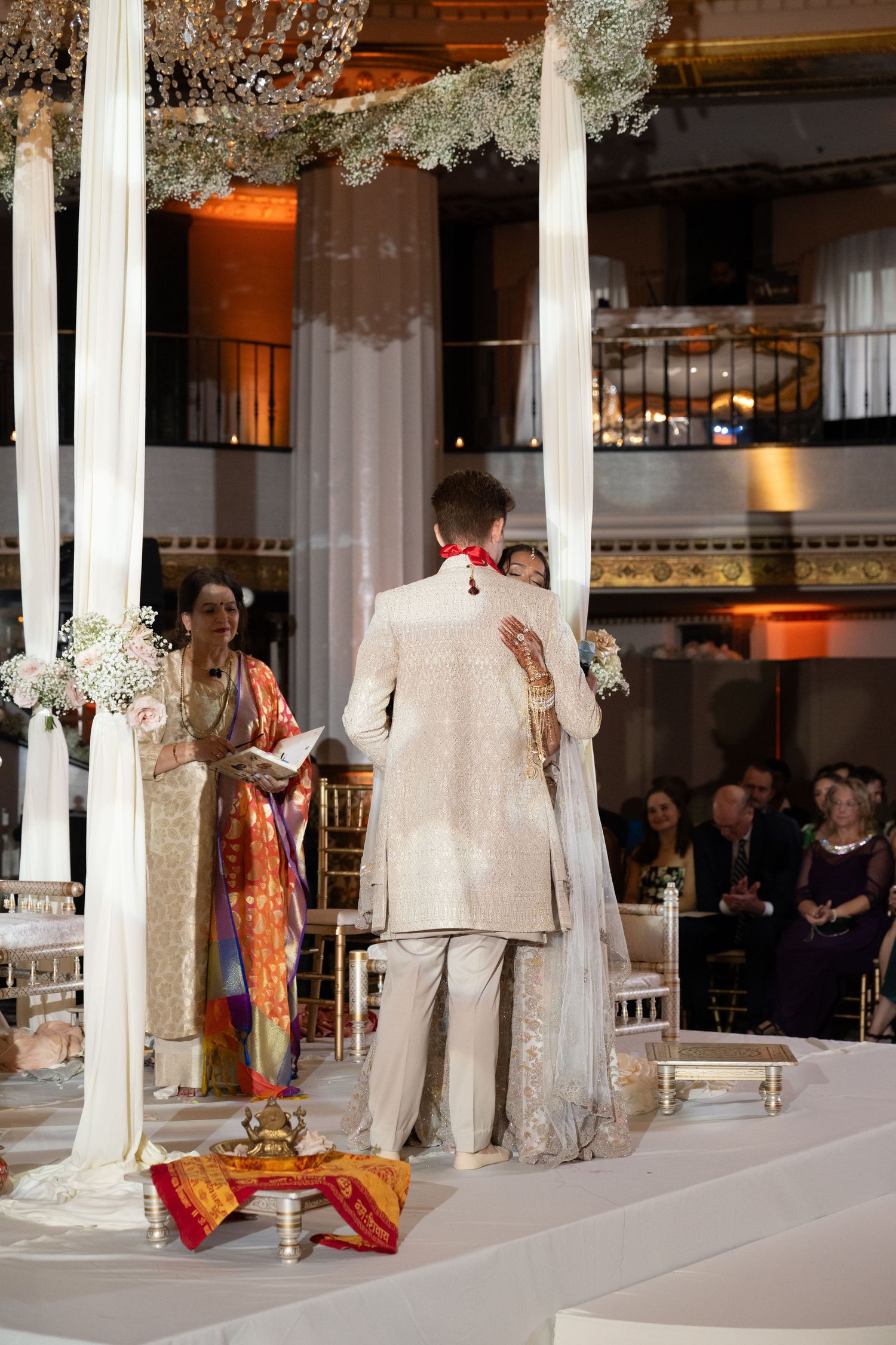 A traditional Indian wedding ceremony takes place in an elegant white-columned venue decorated with floral arrangements.