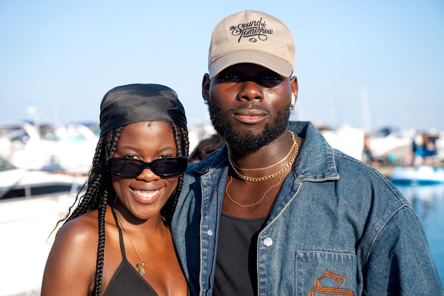 Couple wearing casual attire poses together at a marina with boats in the background.