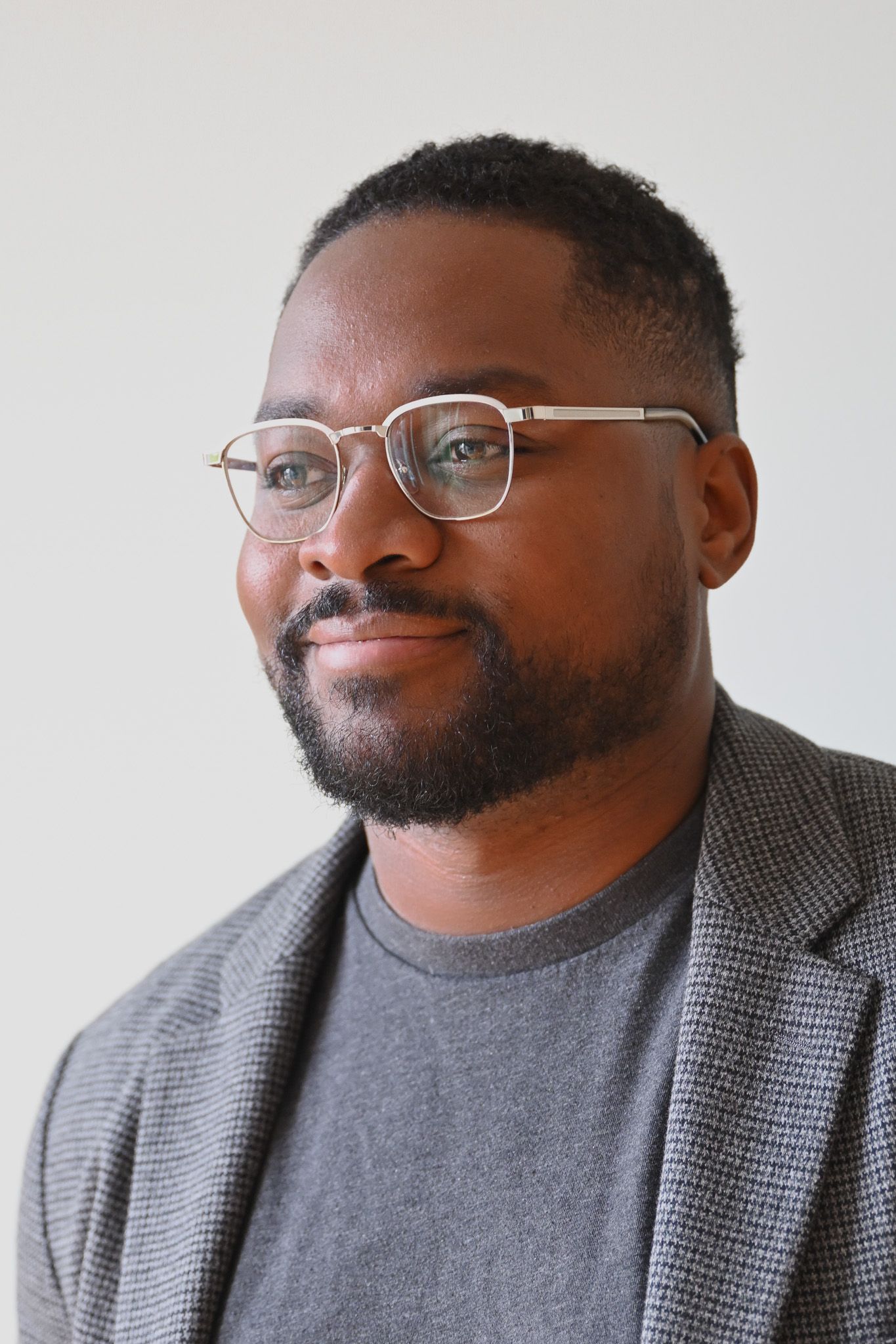 Professional headshot of person in glasses and grey blazer against white wall.