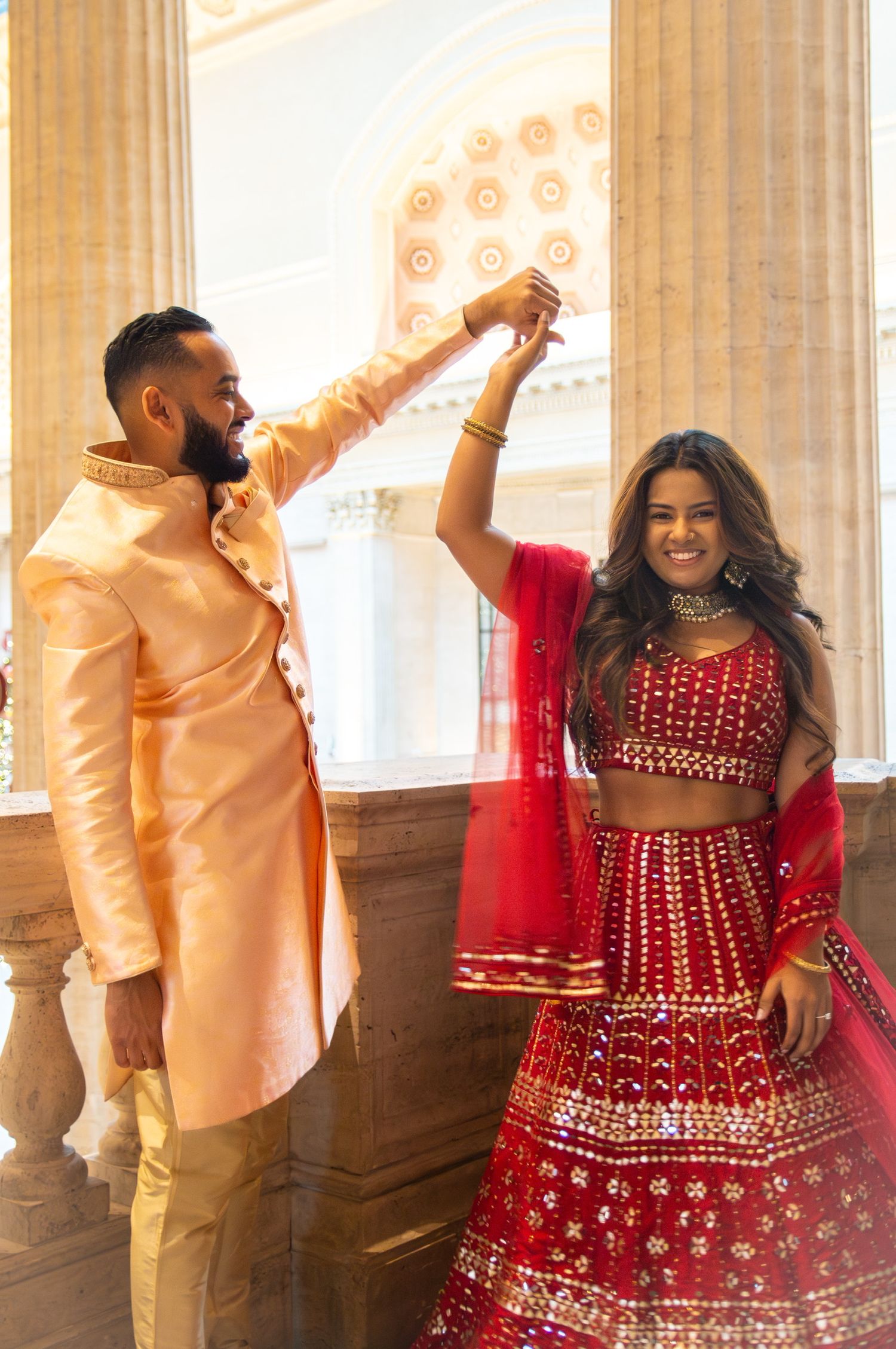 Couple dancing in Indian wedding outfits with bride in red lehenga and groom in peach sherwani.