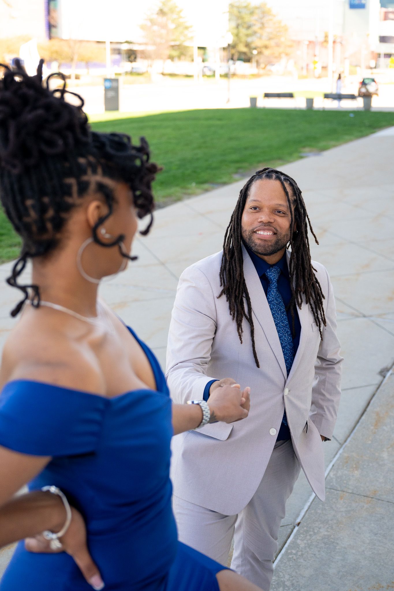 A couple in formal attire smiling and embracing outdoors, with the person in a gray suit and blue shirt.