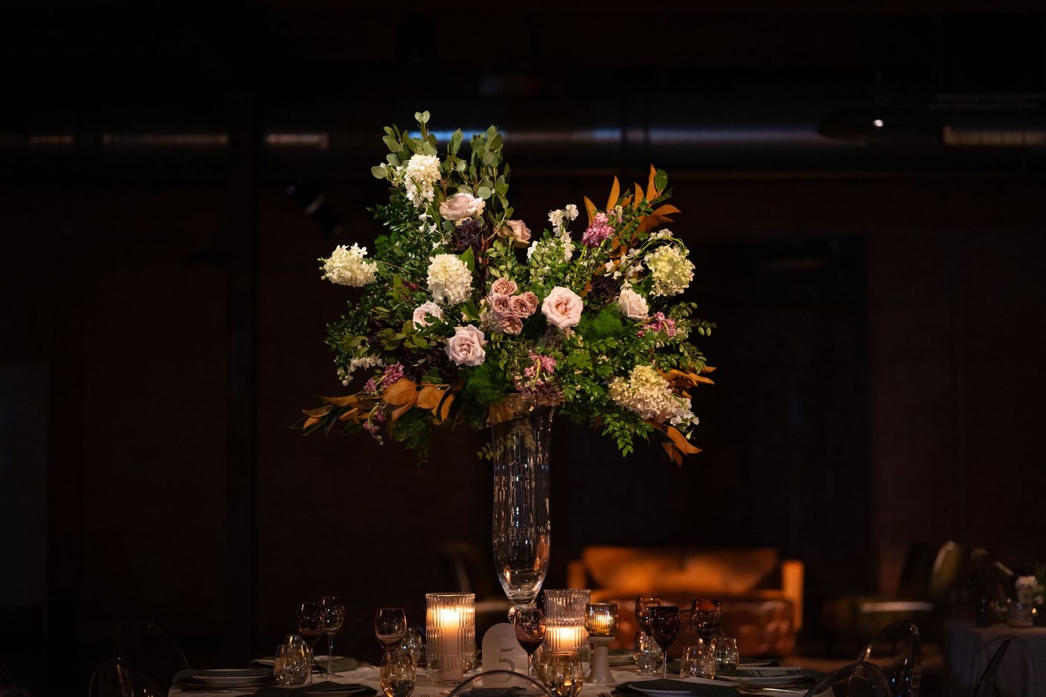 A tall floral centerpiece with white and pink flowers sits atop a dark table with glowing candles in the background.