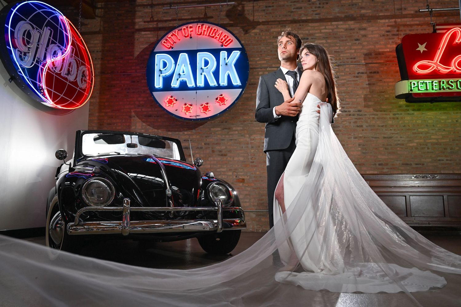 A bride in a flowing white gown poses in front of rustic brick walls with illuminated neon signs in the background.