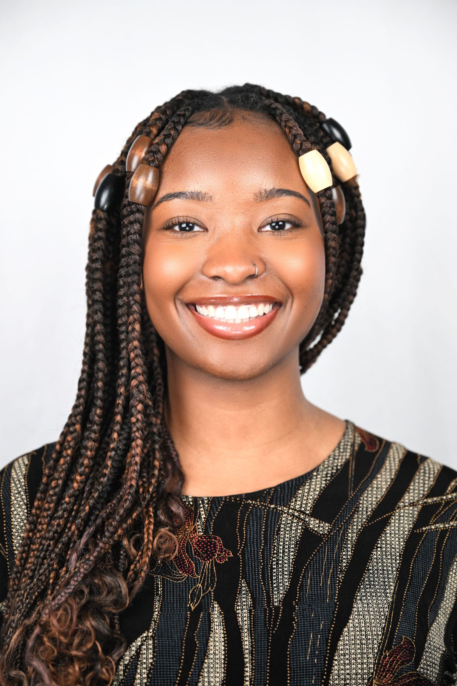 Portrait of someone with long braids decorated with beads wearing a patterned top against white background.