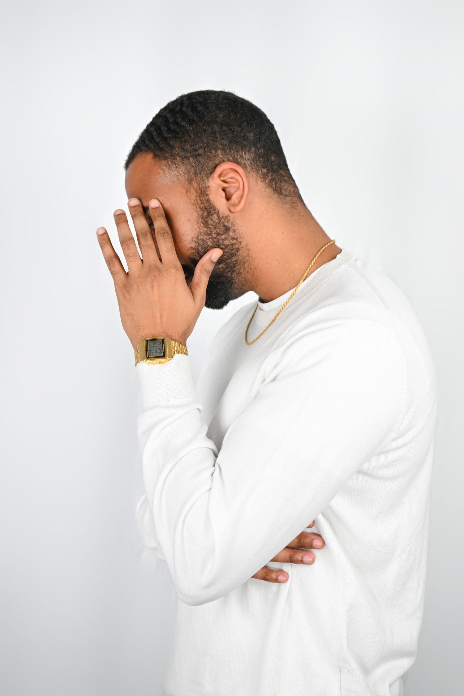 Person wearing a white sweater and gold watch covering face with hands against light background.