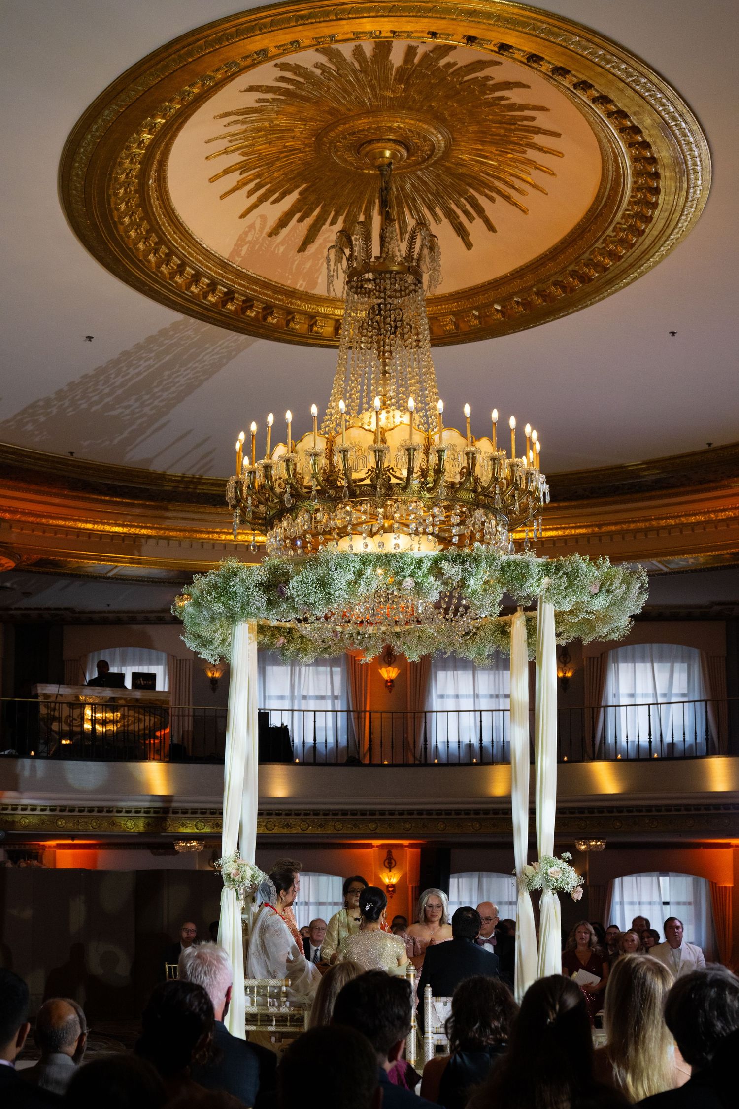 Elegant ballroom with ornate chandeliers and decorative ceiling medallions showcases wedding reception setup with floral arrangements.
