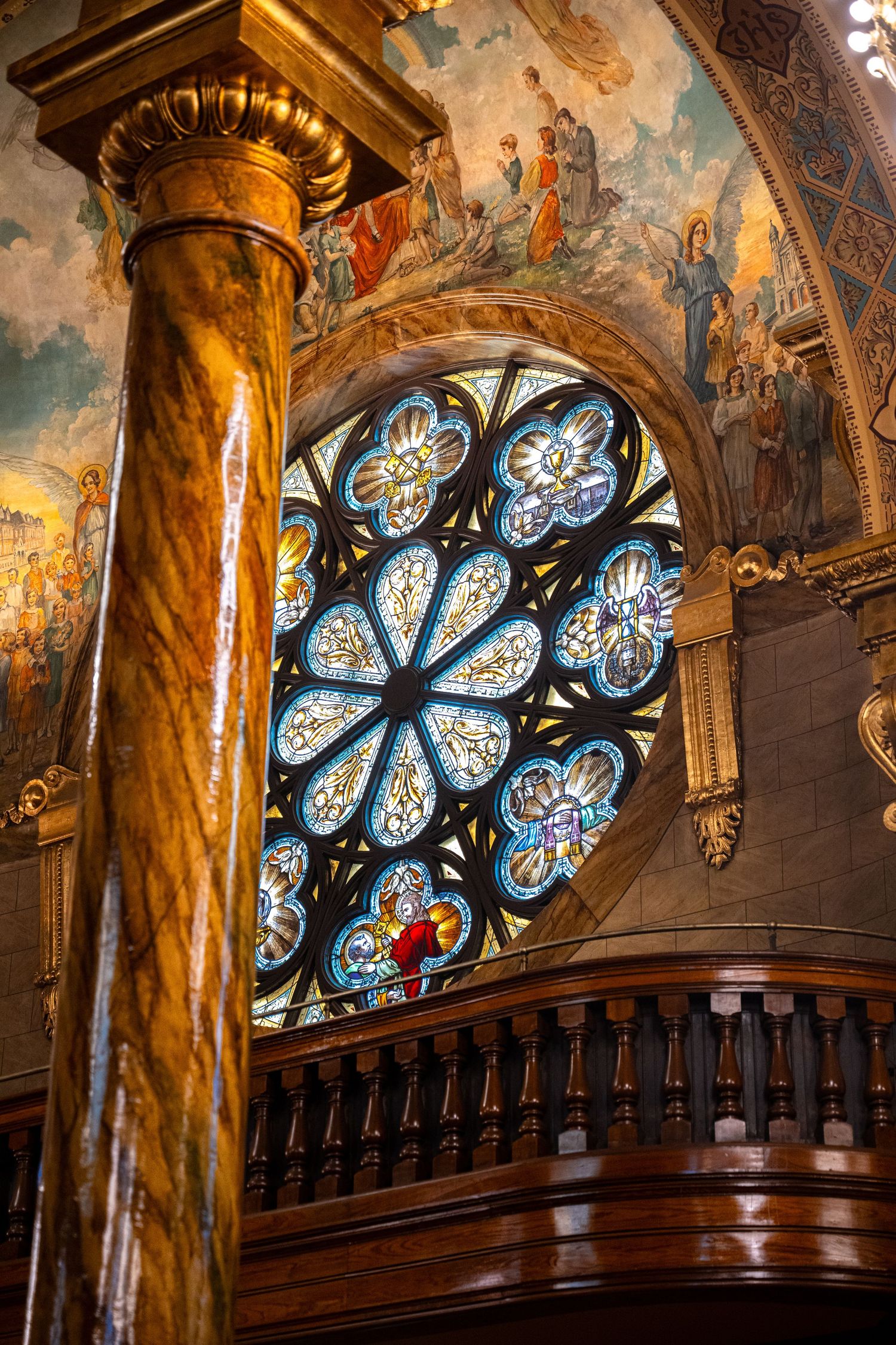 Ornate stained glass rose windows illuminate the interior of a historic church with golden columns and dark wood railings.