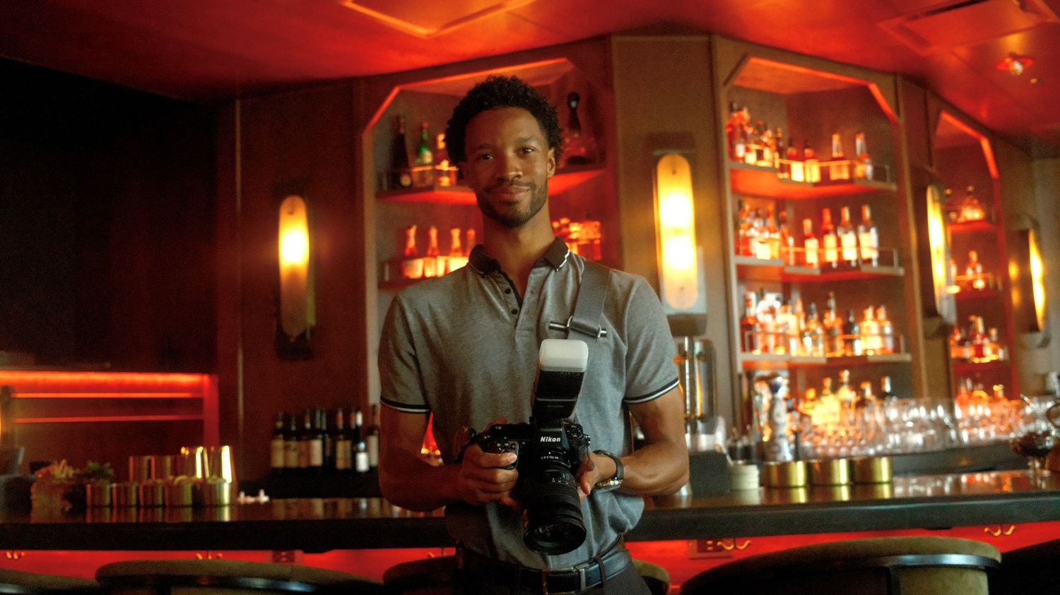 A bartender stands behind a bar counter with red ambient lighting and shelves of liquor bottles in the background.