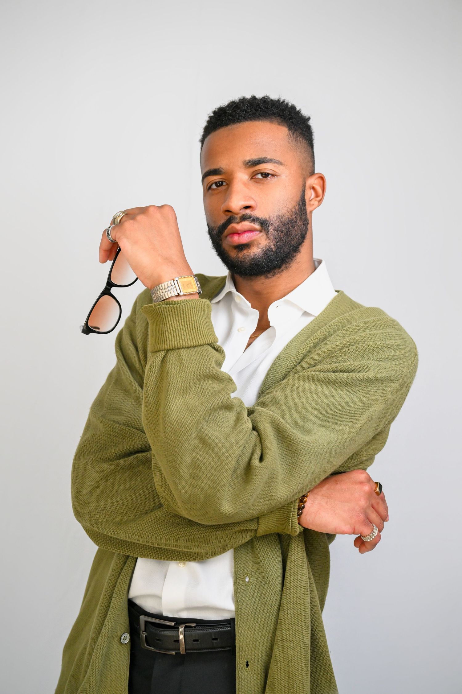 A person in an olive green cardigan and white shirt poses expressively against a light gray background.