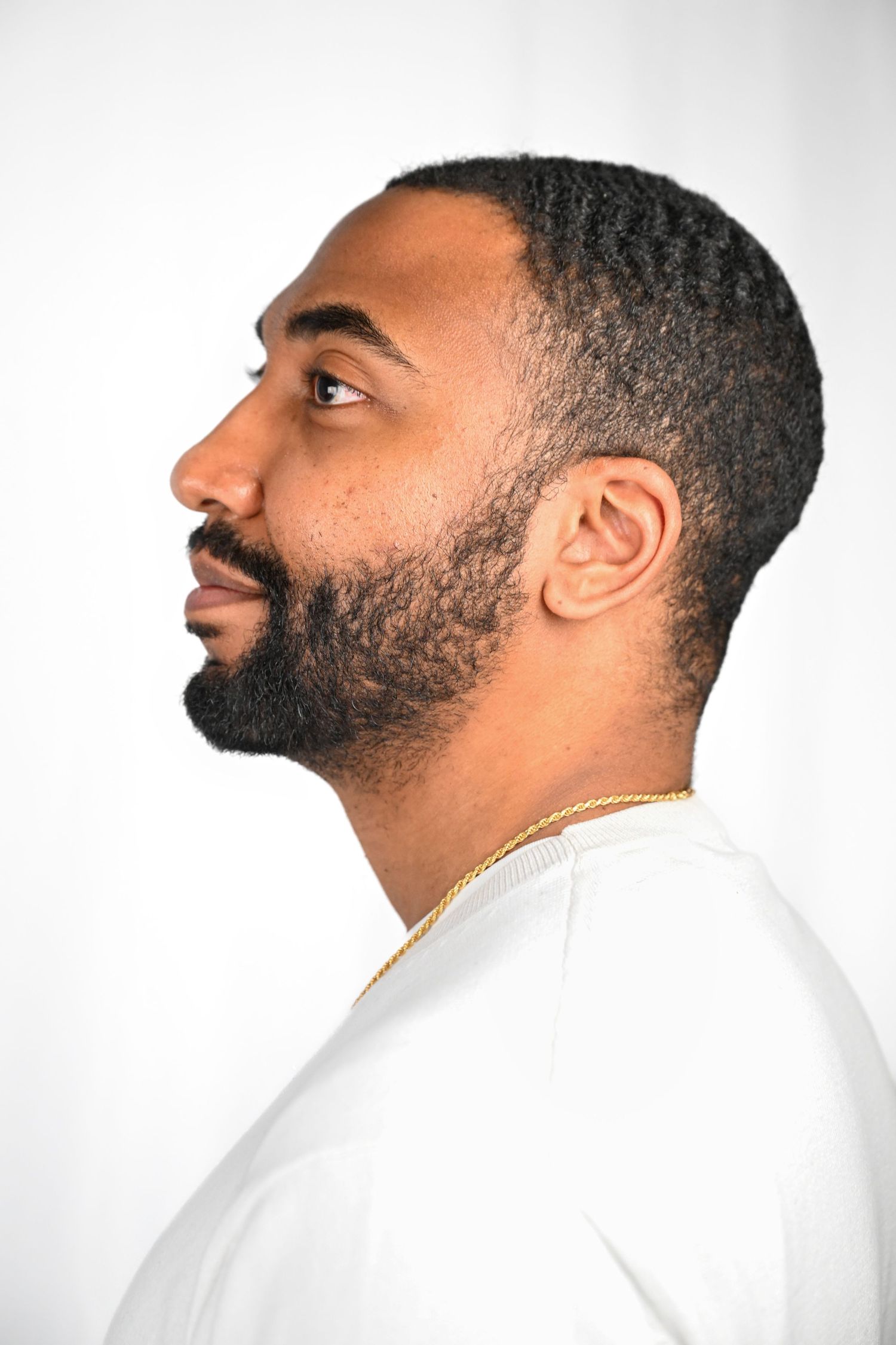 Side profile portrait of person in white top with neatly trimmed beard and gold chain.