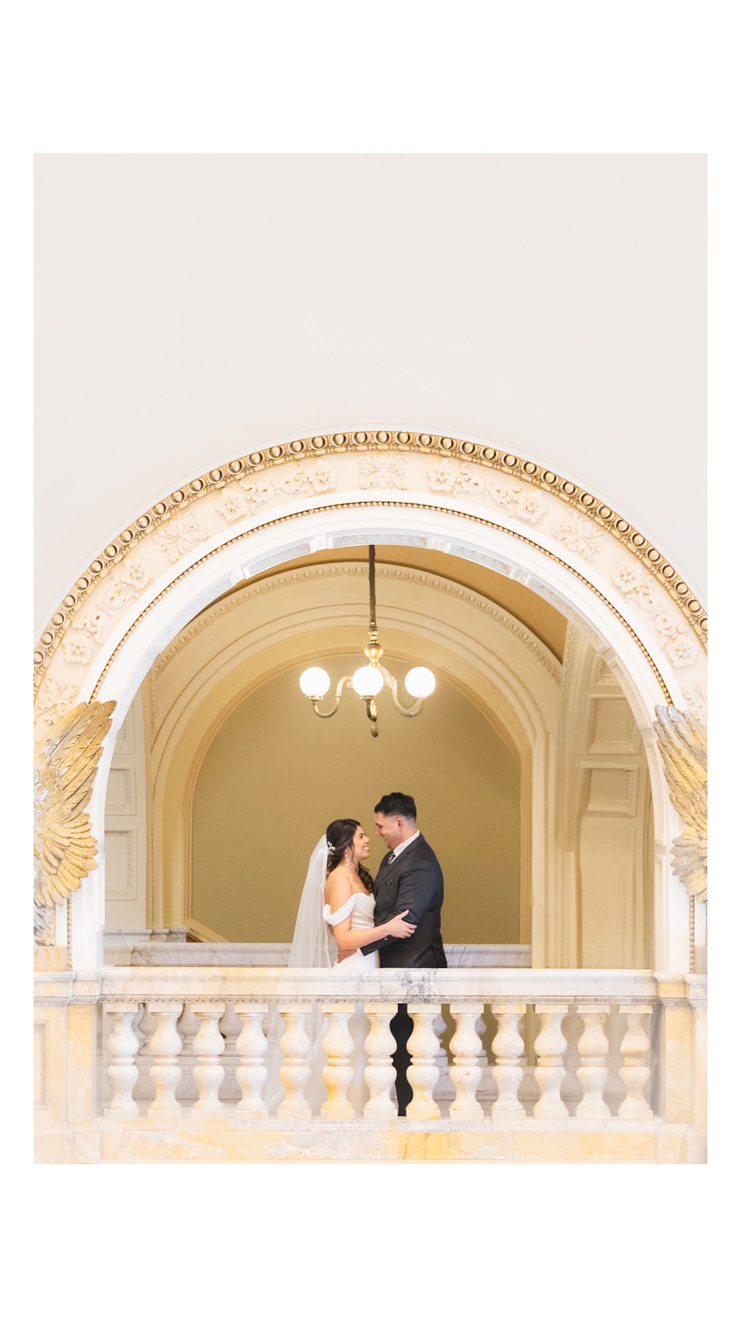 A romantic wedding couple embraces under an ornate cream-colored archway with a chandelier.