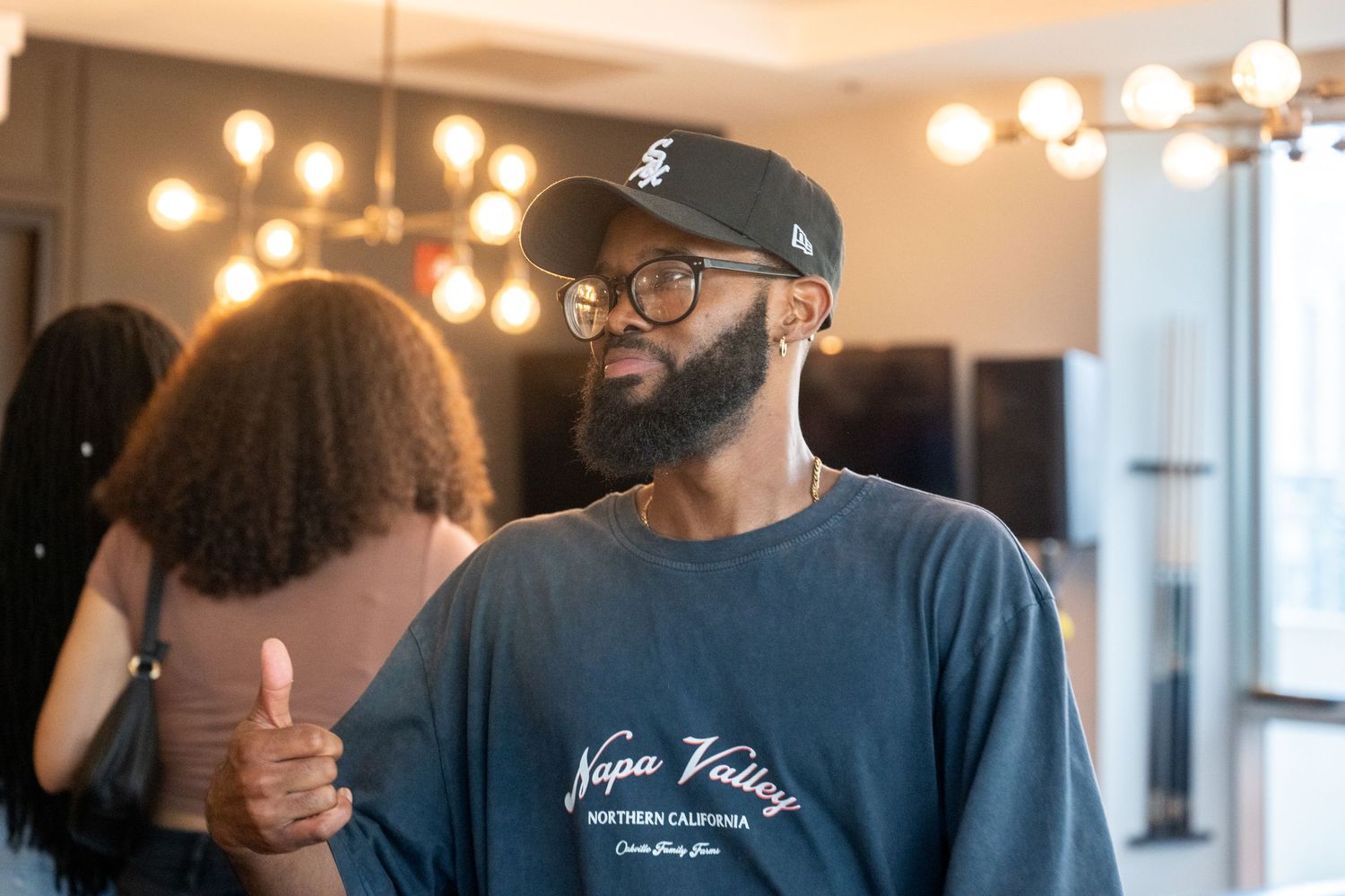 Person wearing baseball cap and glasses giving thumbs up in warmly lit room.