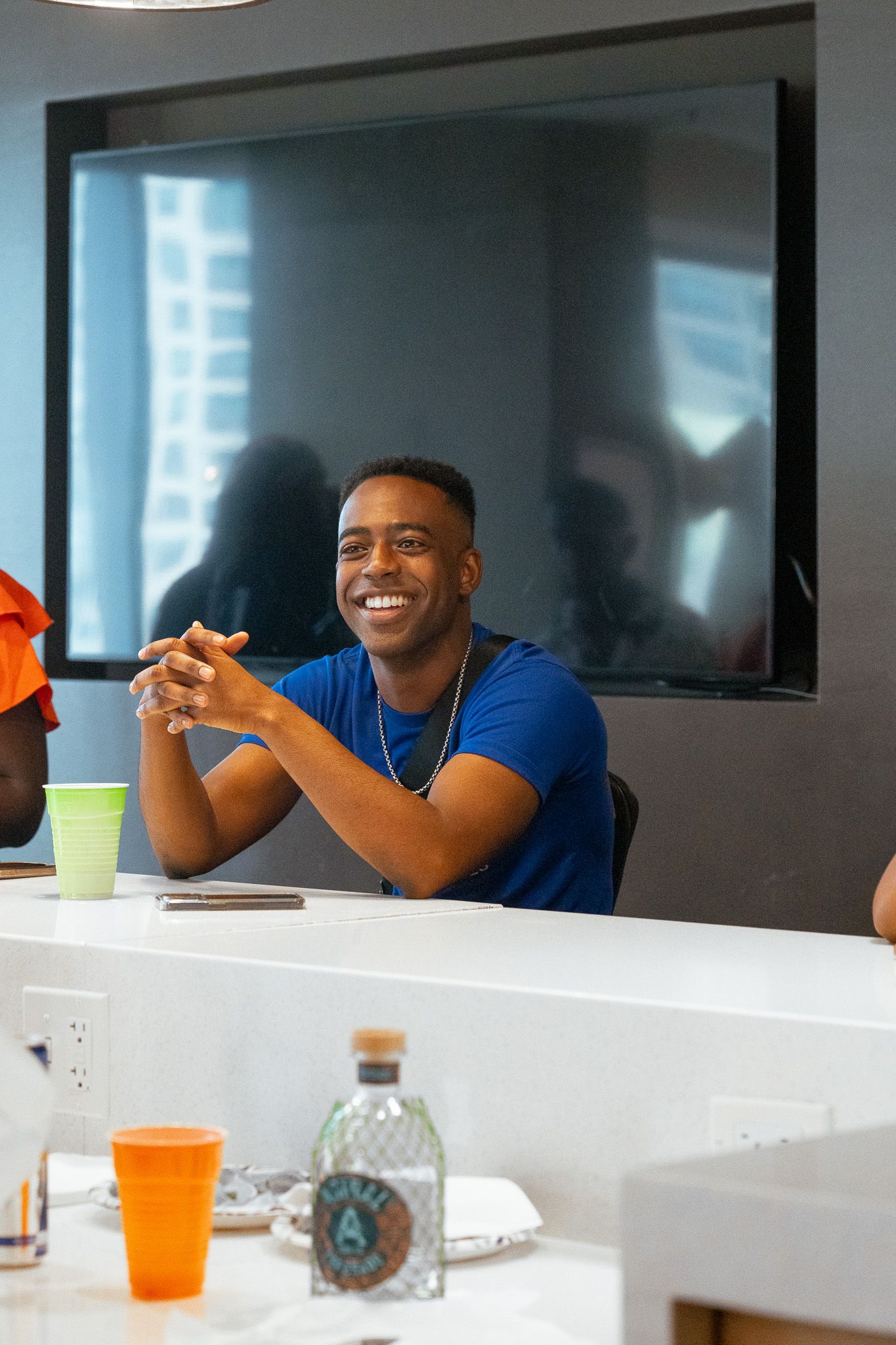 Person in blue shirt smiles while sitting at conference table with drinks.