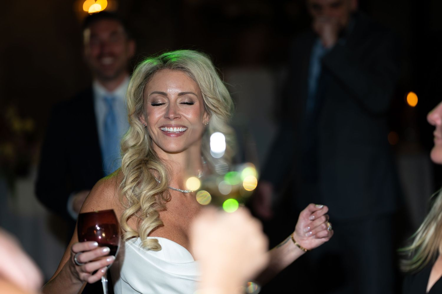 Woman in a white dress laughs while holding a drink at a dimly lit party or event.