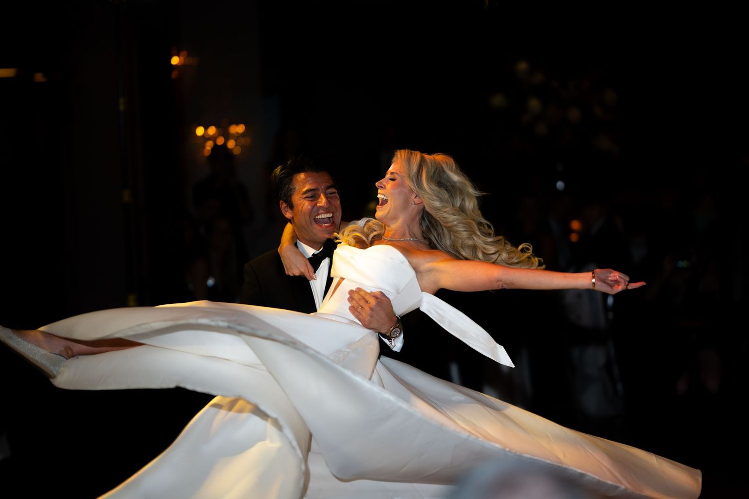 A couple in elegant wedding attire performs a graceful dip during their first dance on a dark dance floor.