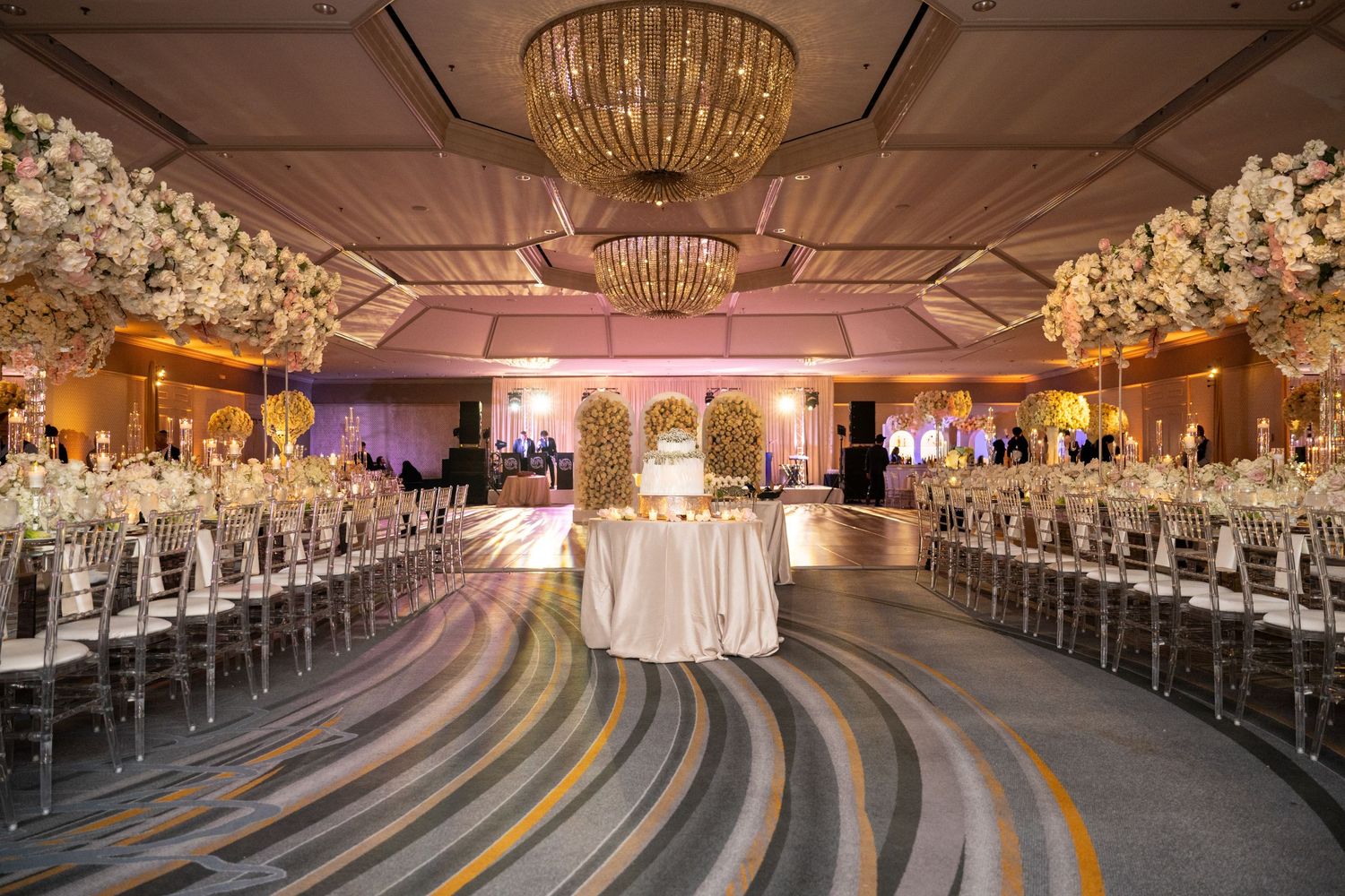 Elegant ballroom with crystal chandeliers, white floral arrangements, and curved rows of chairs set up for a wedding ceremony.