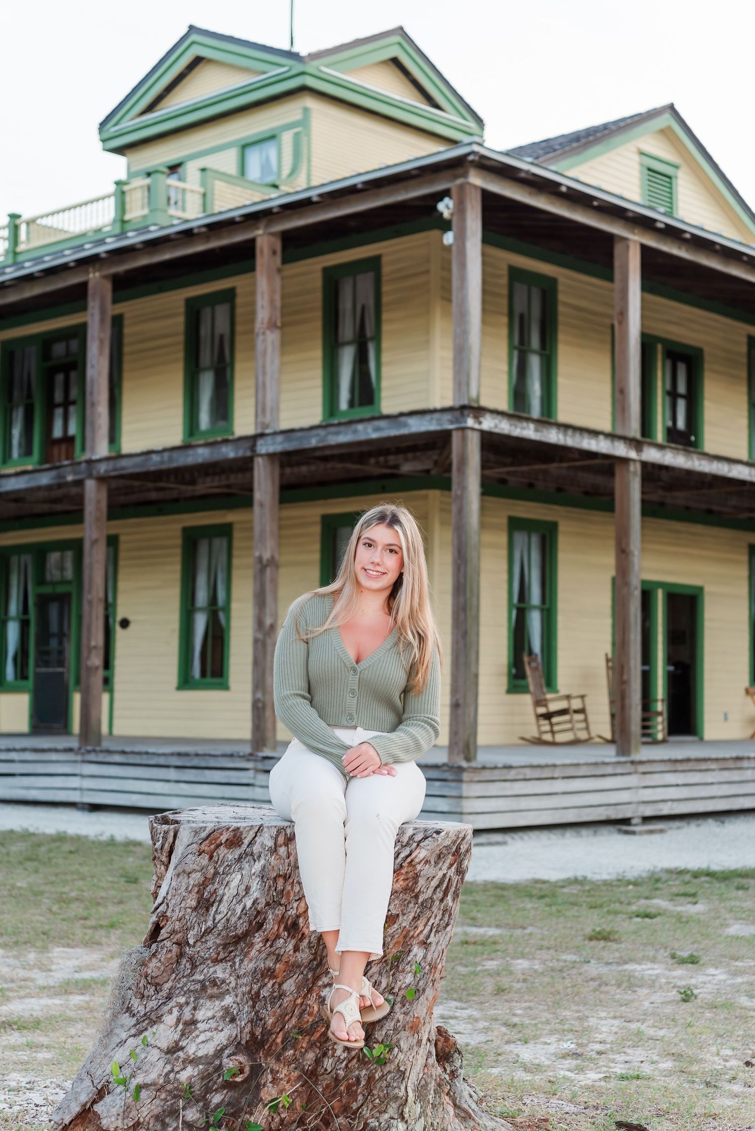 Historic yellow two-story house with wraparound porch and green trim serves as backdrop for casual outdoor portrait.