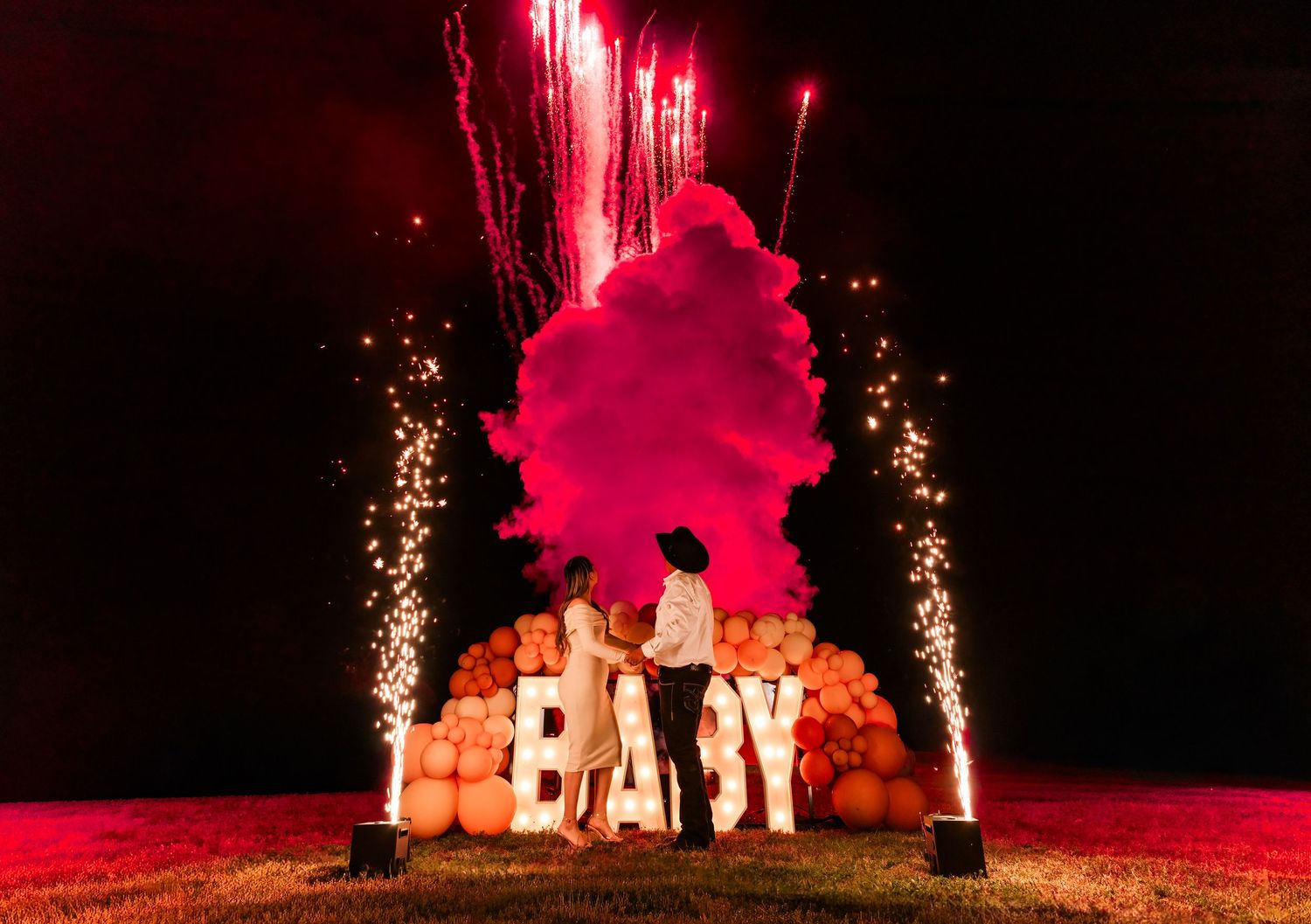 Pink fireworks and sparklers illuminate a 'BABY' sign display against a dark night sky.