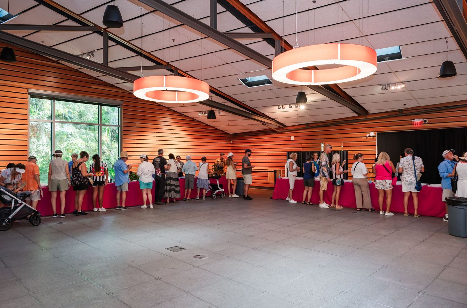 Guests mingle at an indoor event space with warm wood walls, round pendant lights, and red-draped tables.