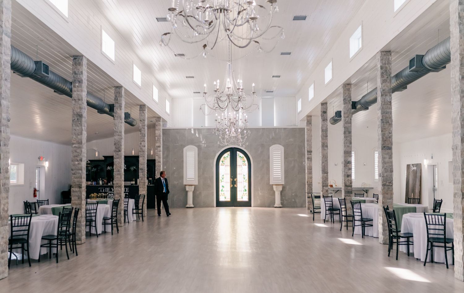 Elegant wedding venue ballroom with chandeliers, brick columns, hardwood floors, and round tables set for an event.