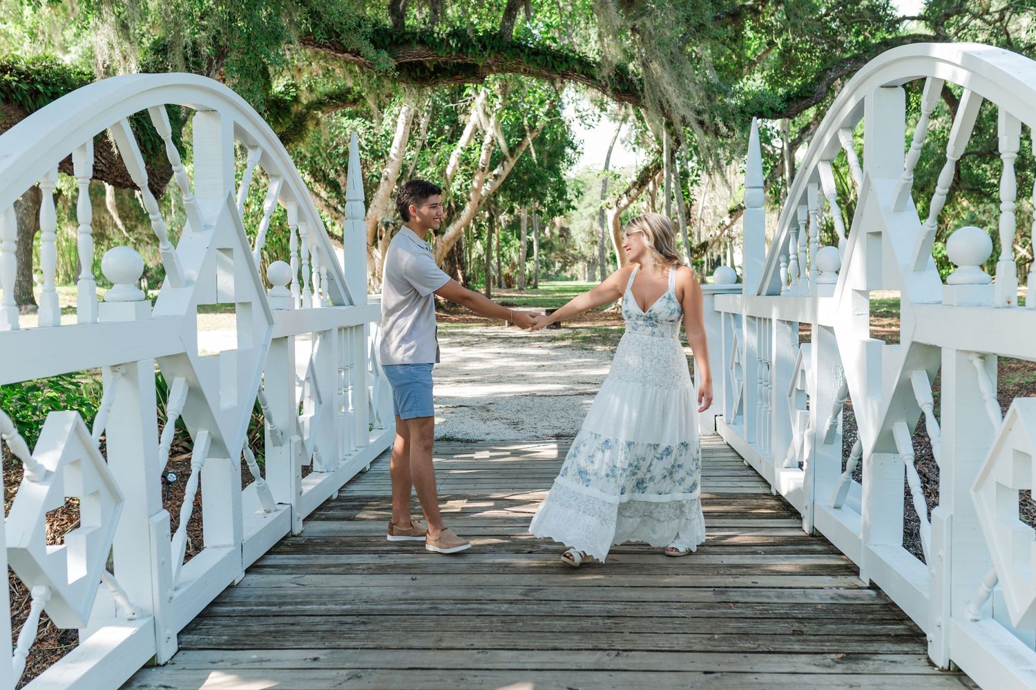 Couple holding hands on a white wooden bridge surrounded by lush green trees in an outdoor park setting.
