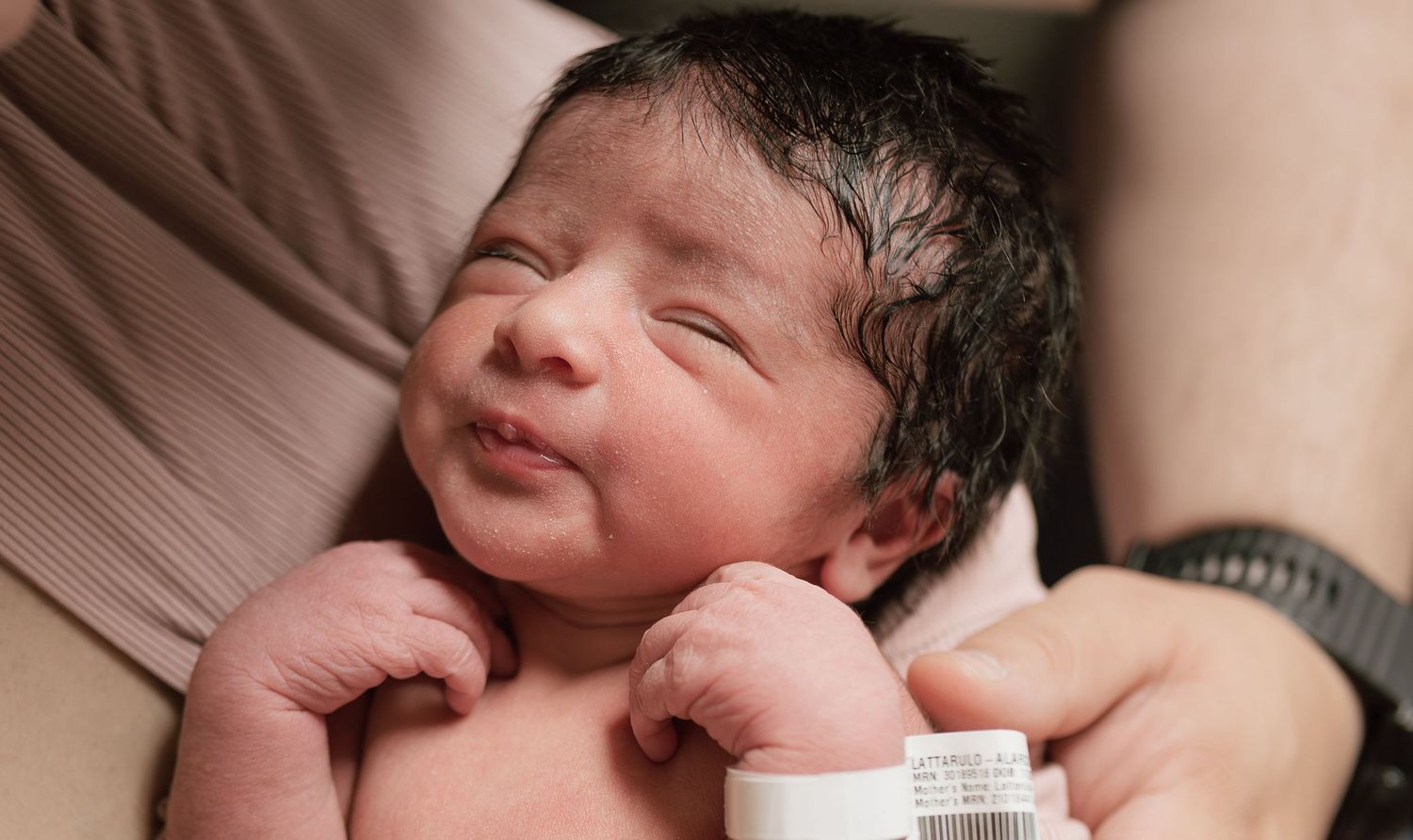 Newborn baby with dark hair smiling peacefully while being held, wearing a hospital identification bracelet.