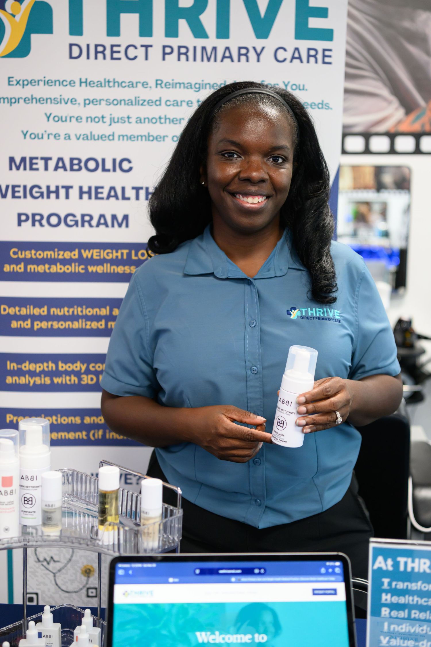A smiling woman in a blue Thrive Direct Primary Care shirt holds a product at a metabolic weight health program booth.
