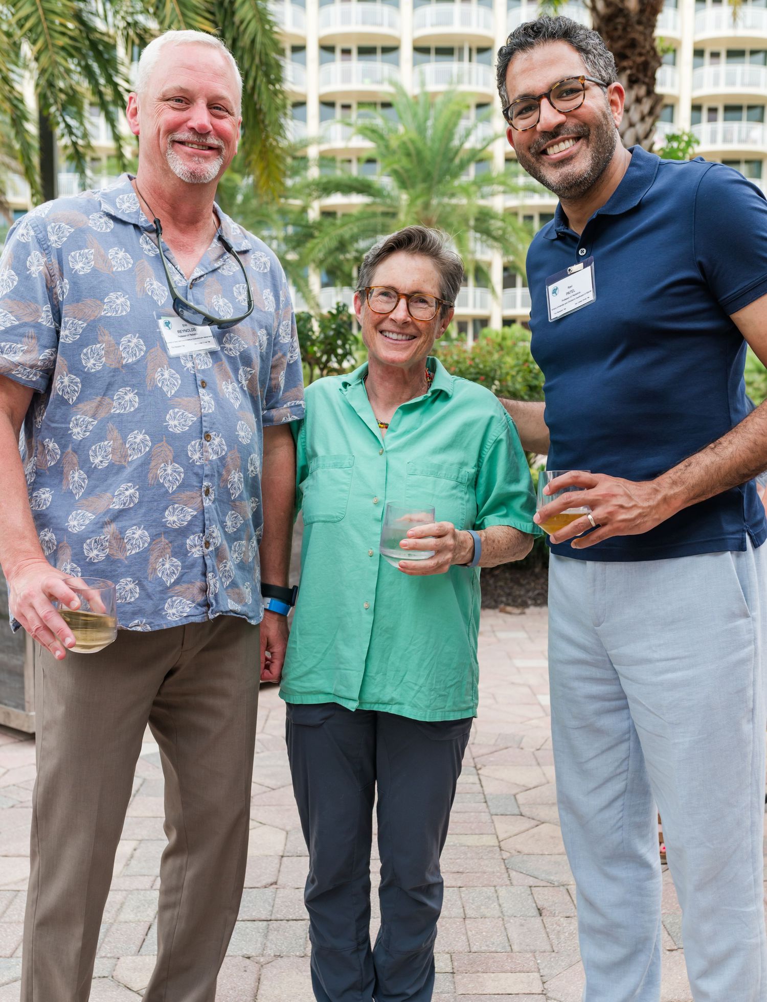 Three smiling attendees wearing name tags enjoying drinks at an outdoor tropical resort event.