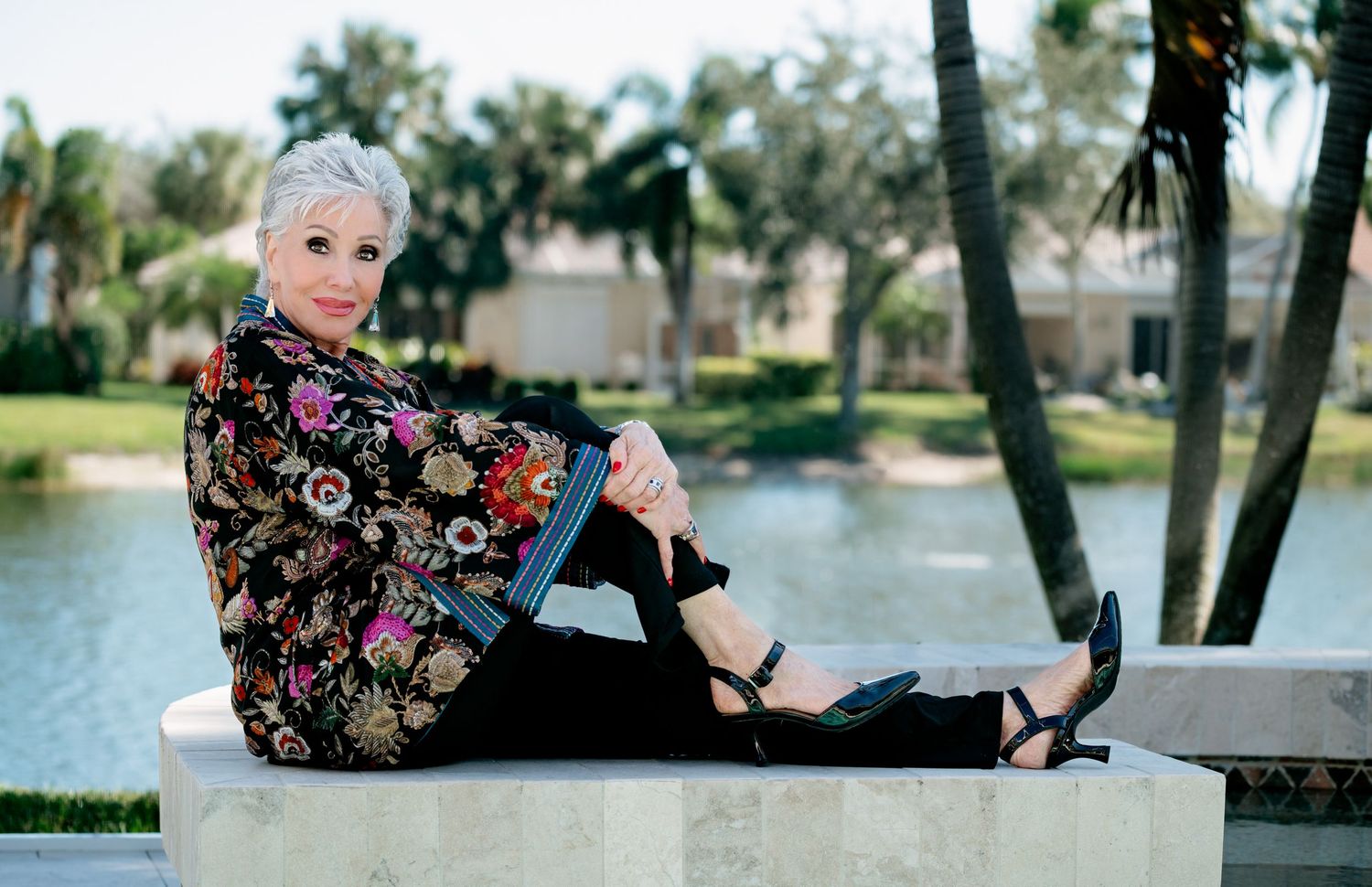 Elegant older woman with short white hair sits outdoors in floral jacket and black pants near a serene Florida waterway.