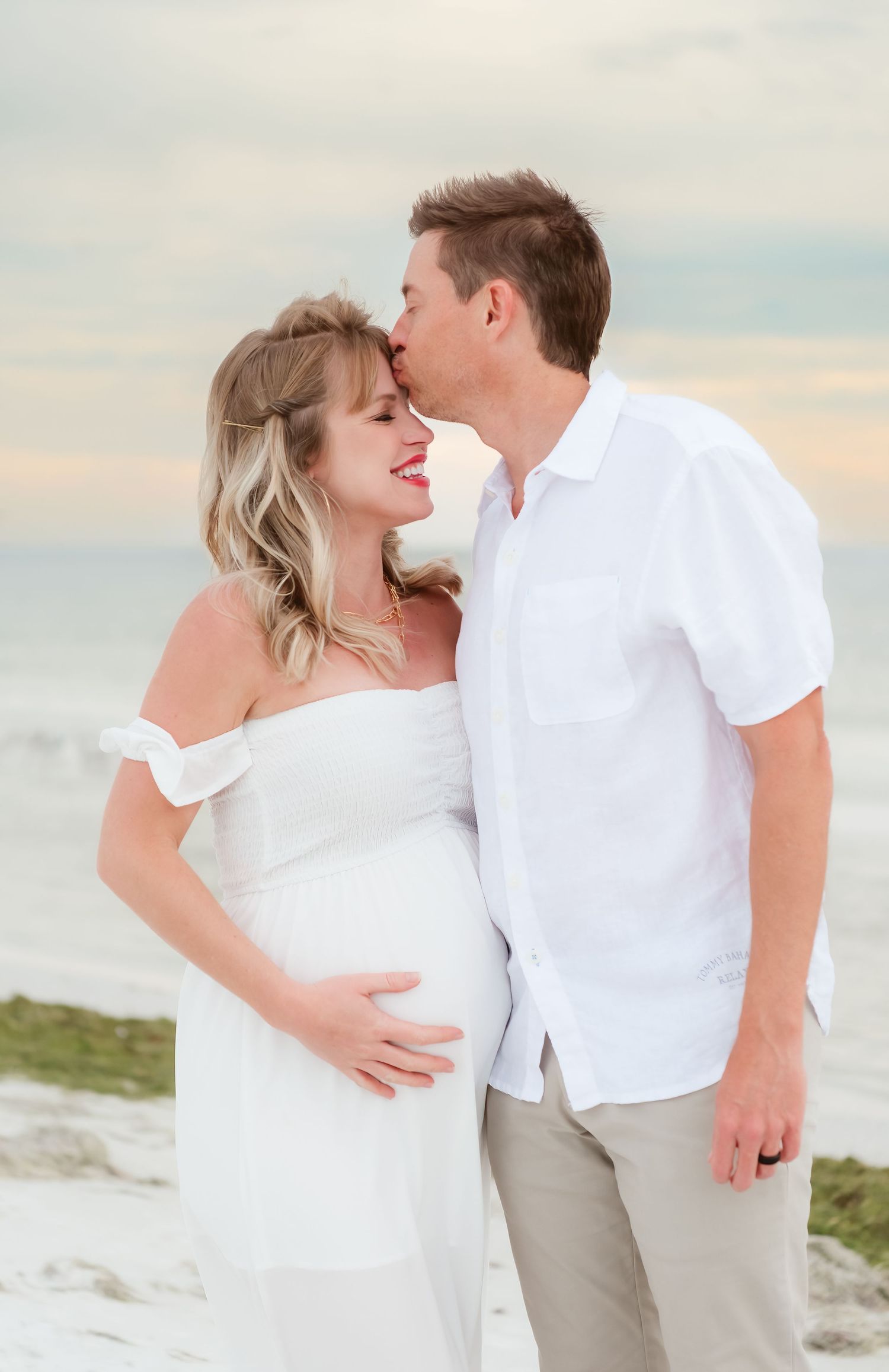 Couple in white outfits sharing a tender kiss on a sandy beach, with the woman cradling her pregnant belly.