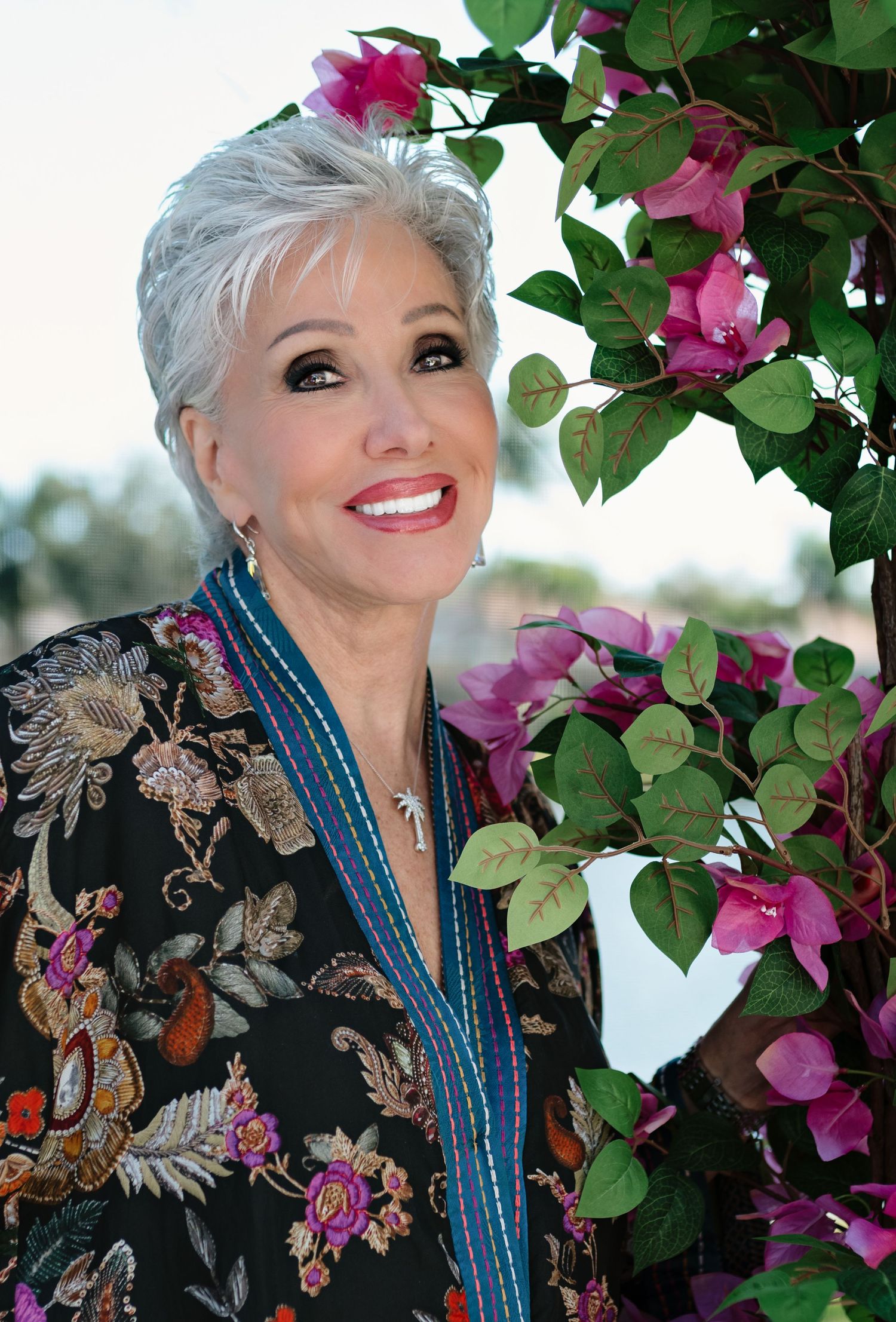 Elegant older woman with short gray hair smiling beside pink bougainvillea, wearing a floral patterned black jacket.