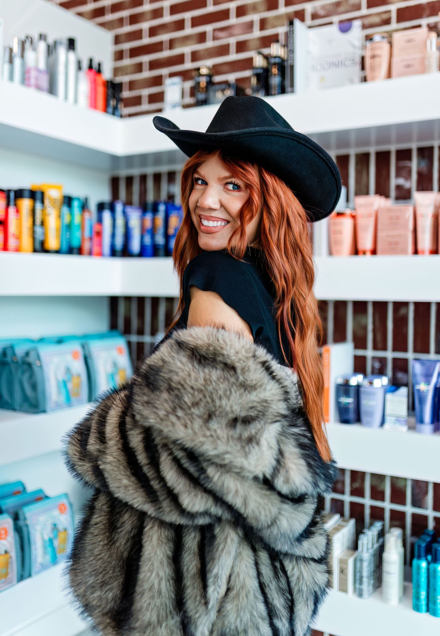 Person wearing a black baseball cap and grey fur coat poses against a brick wall with shelves of colorful books.