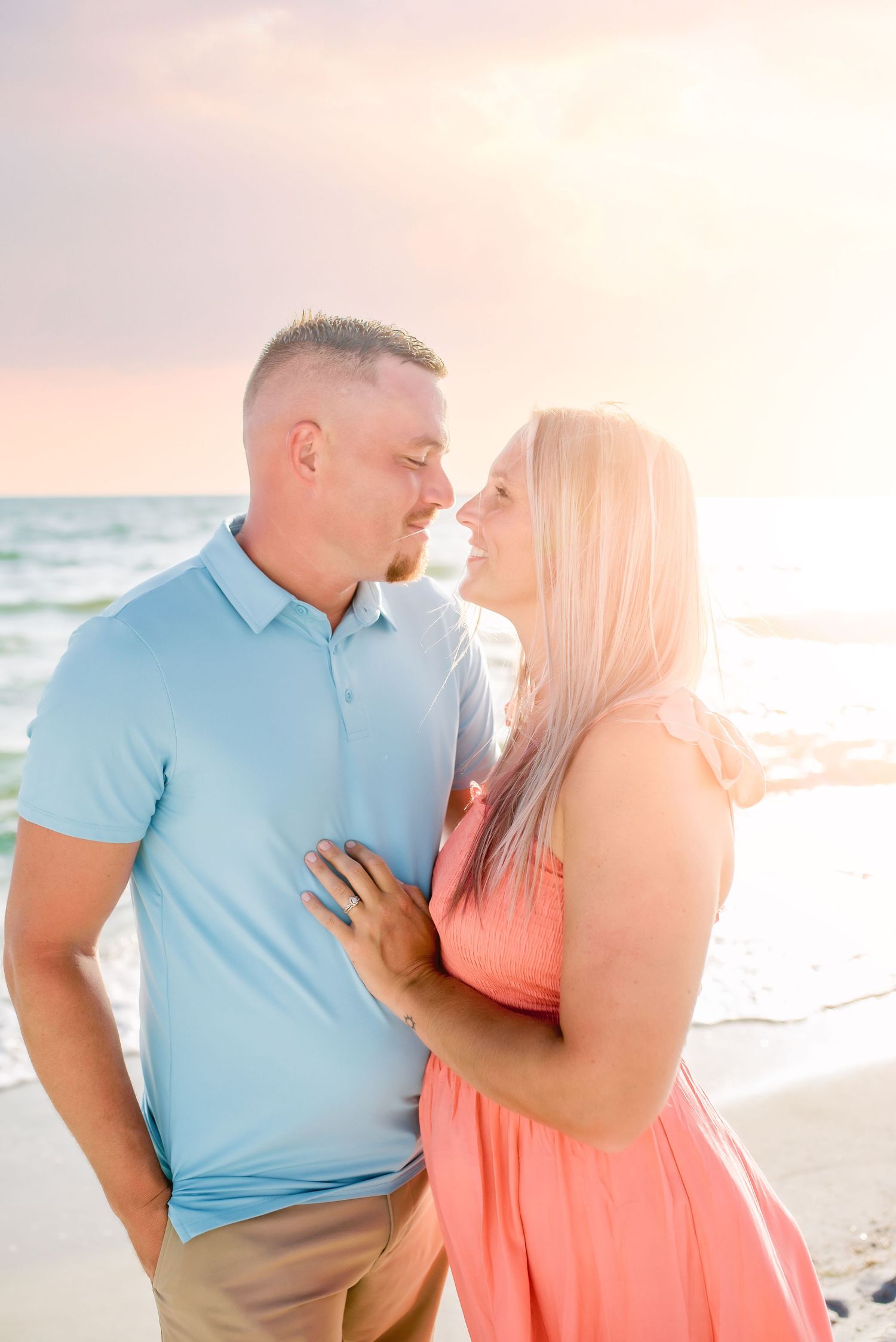A couple in blue and coral outfits share romantic moments on a sunlit beach during a photo session.