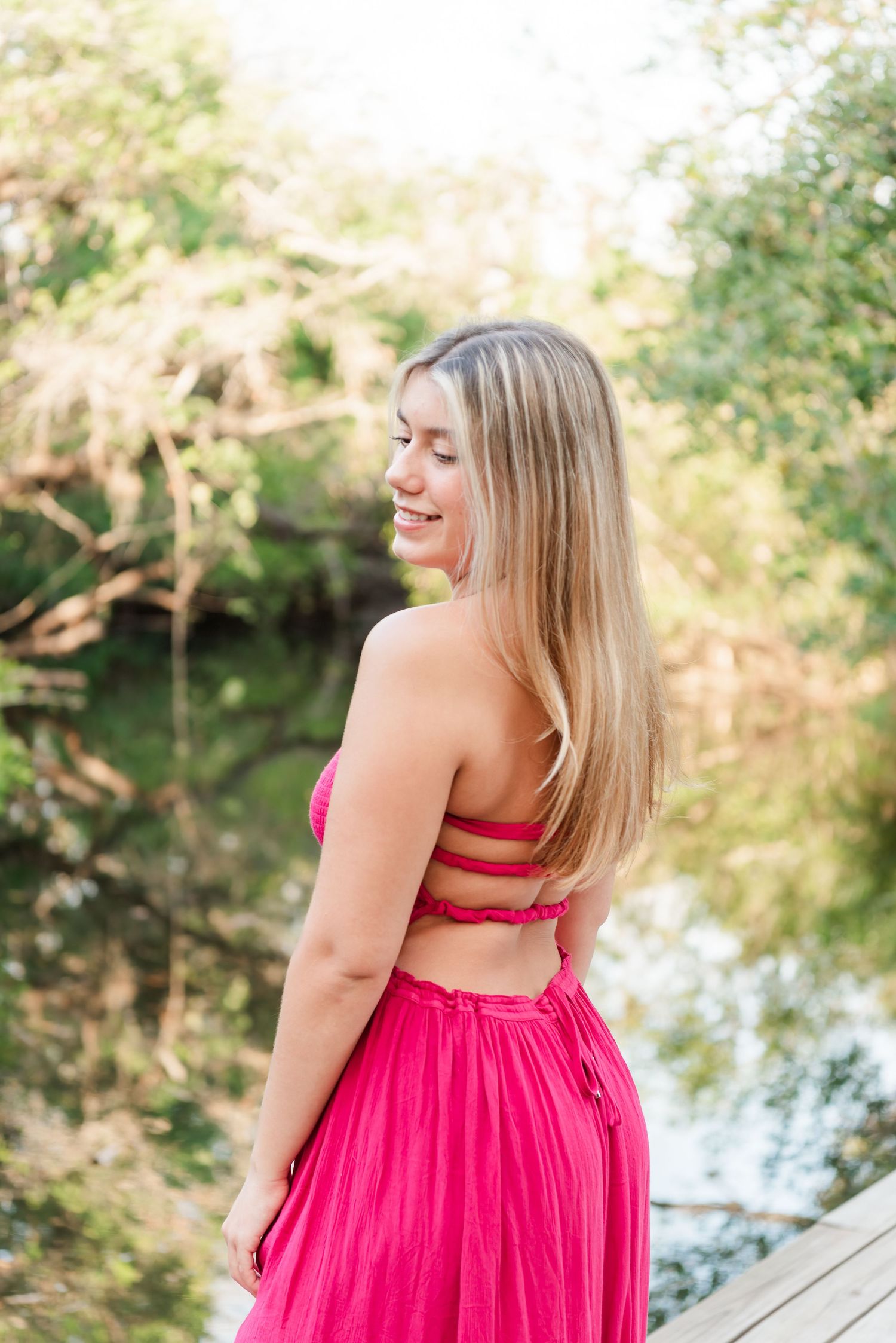 Person wearing a bright pink backless dress poses on a wooden deck by a lake during golden hour lighting.