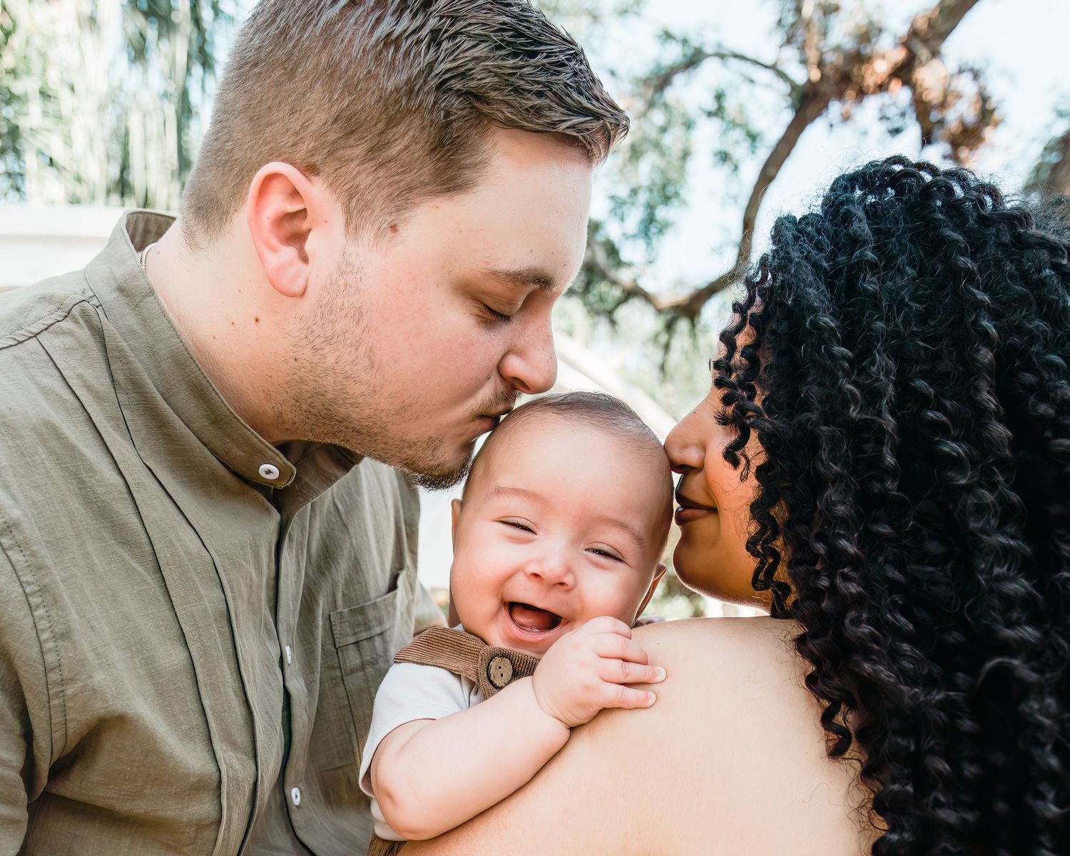A laughing baby shares a joyful moment with loving parents outdoors in a sunlit setting.