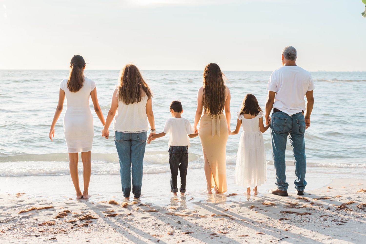 A family wearing coordinated white and blue outfits stands together facing the ocean at sunset on the beach.