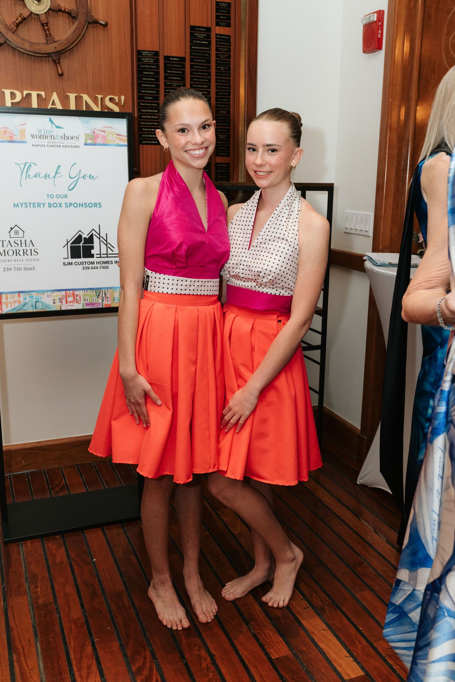 Two people in matching coral-colored dresses pose together on hardwood floors near nautical decor.