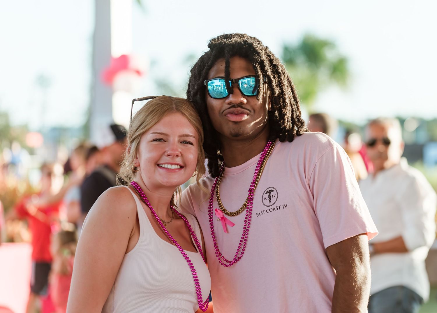 Two festival attendees pose together outdoors wearing sunglasses and beaded necklaces at a sunny summer event.
