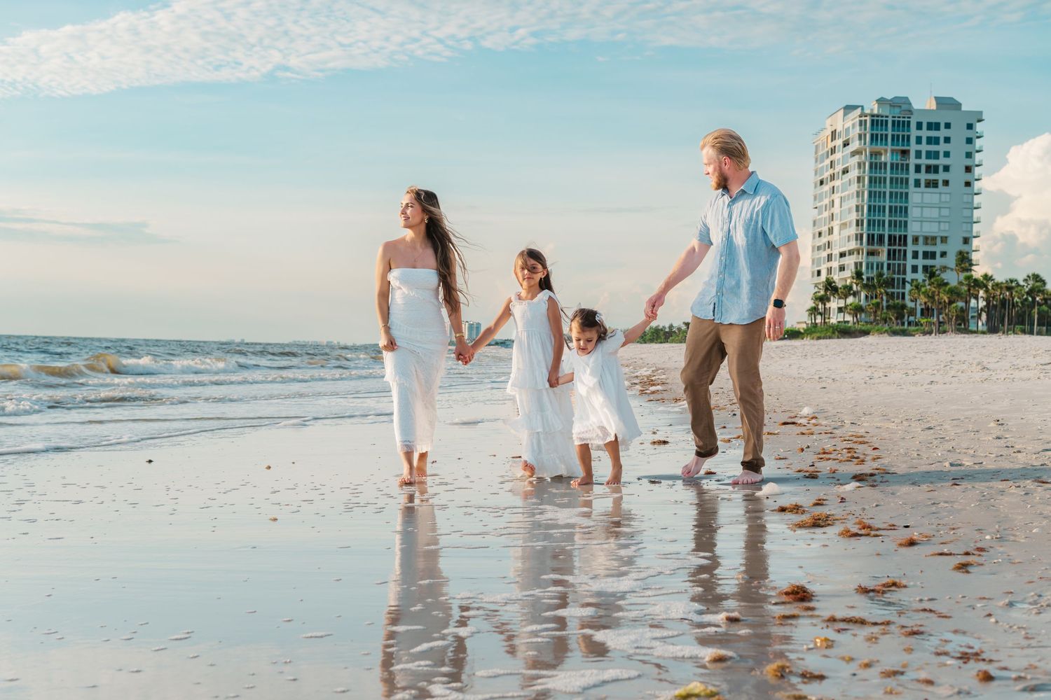 Family of four holding hands and walking on a sunny beach with white outfits and a resort in the background.