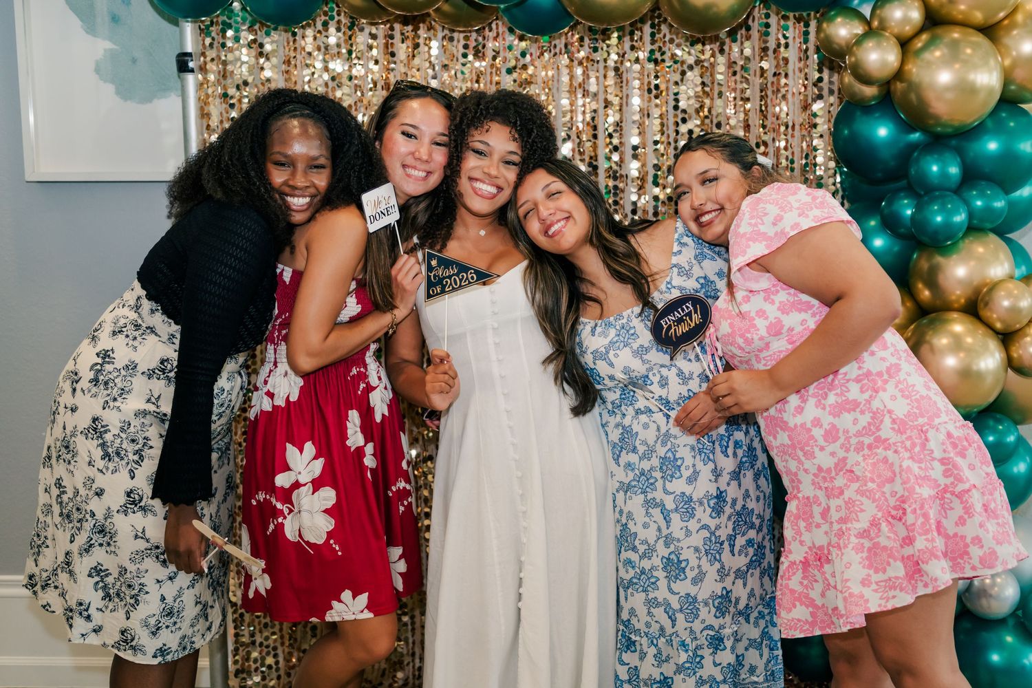 Five women smile together at a celebration event with gold and teal balloon backdrop, two holding photo booth props.