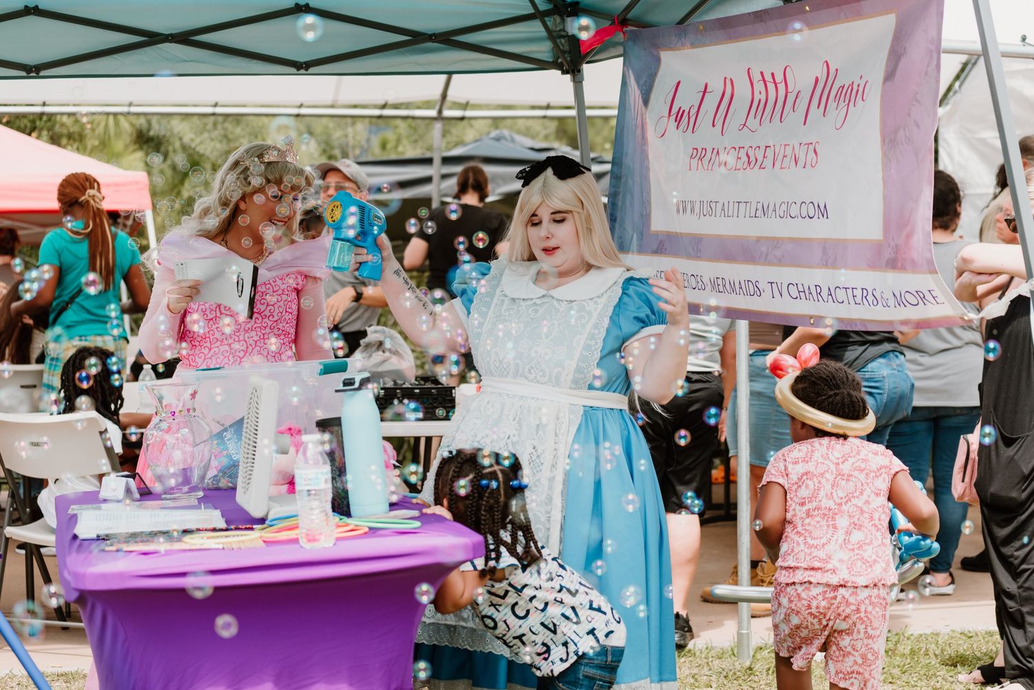 A festive vendor booth at an outdoor market displays pink and purple merchandise with a decorative banner.
