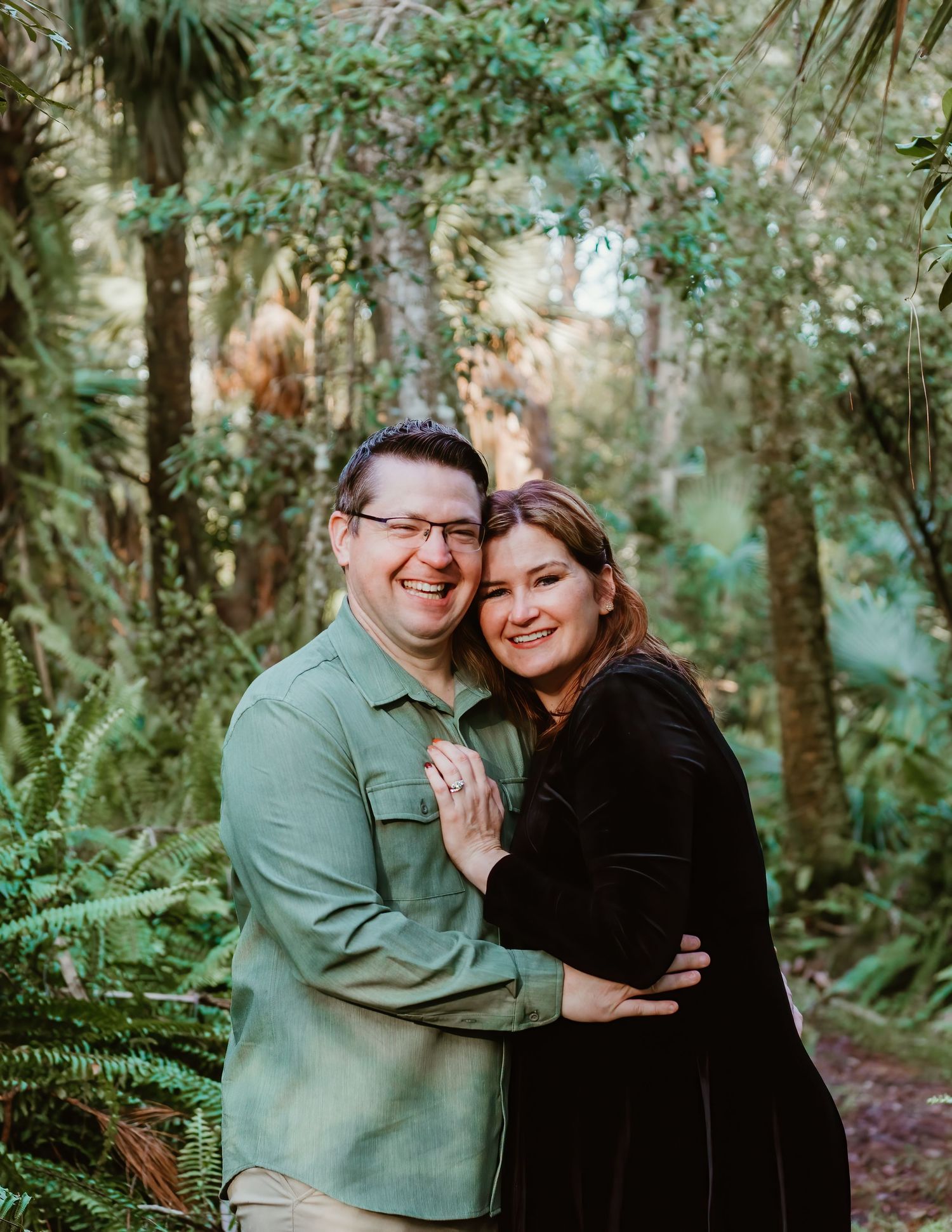 A couple embraces and laughs together while standing in a lush green forest setting.