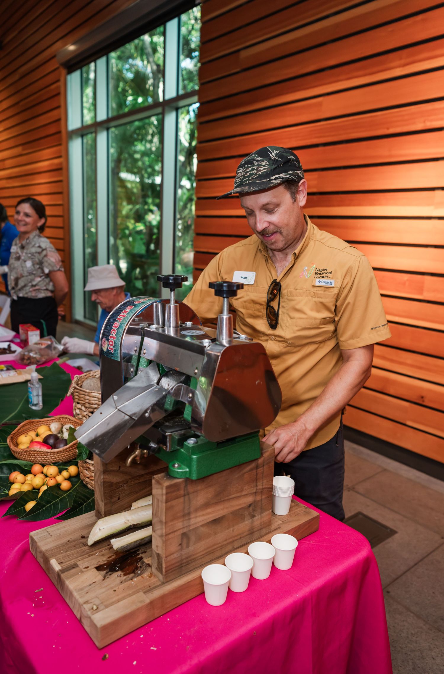 Man in yellow shirt operating a sugarcane juice press machine at an indoor food event with white cups.