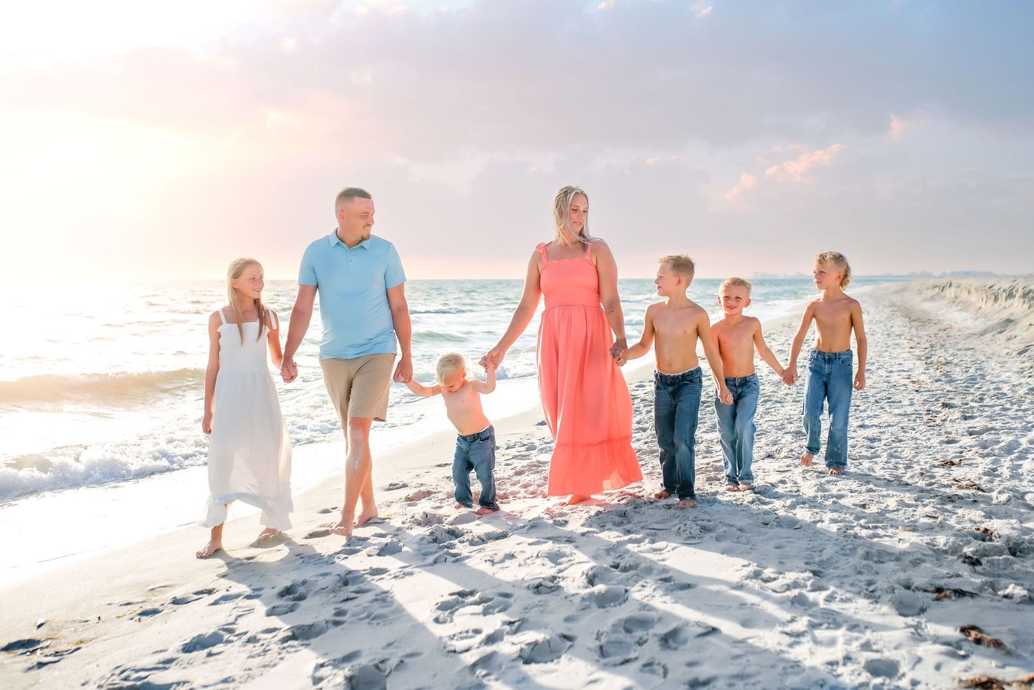 A group of people walking together on a white sandy beach at sunset, holding hands in a line formation.